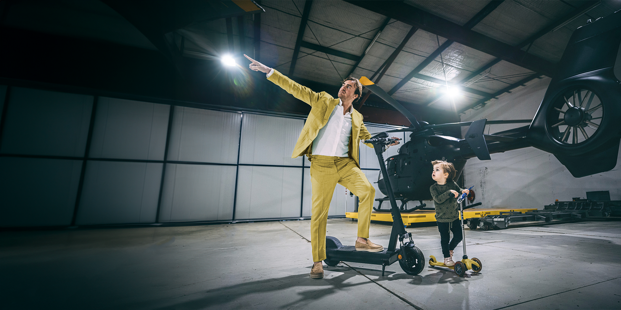 Afbeelding van de Selana e scooter waar een man in een geel pak op staat. Een jongen staat ernaast te kijken. Op de achtergrond staat een zwarte helikopter.