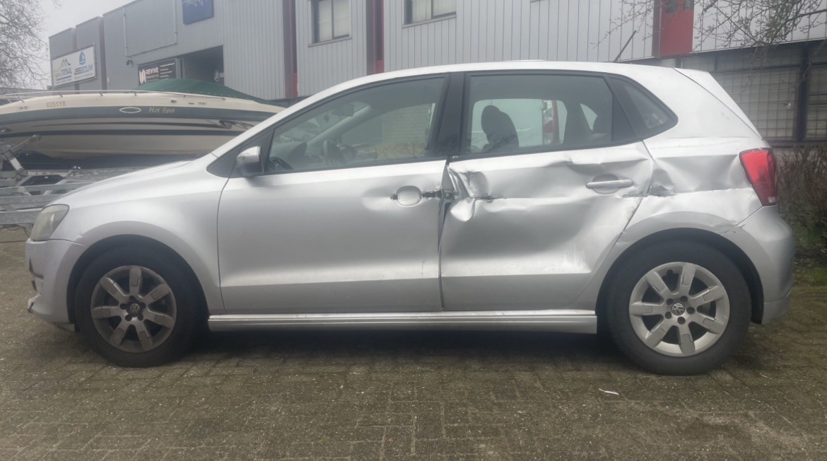 Een zilvergrijze Volkswagen met zware schade aan de rechterzijde, waarbij beide passagiersdeuren en het achterscherm zijn ingedeukt.