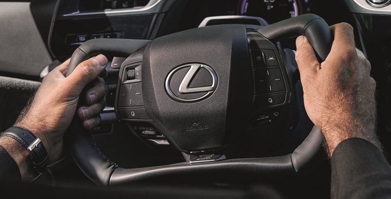 Een close‑up van handen die een modern Lexus‑stuur vasthouden, met een plat boven‑ en onderstuk en diverse bedieningstoetsen.
