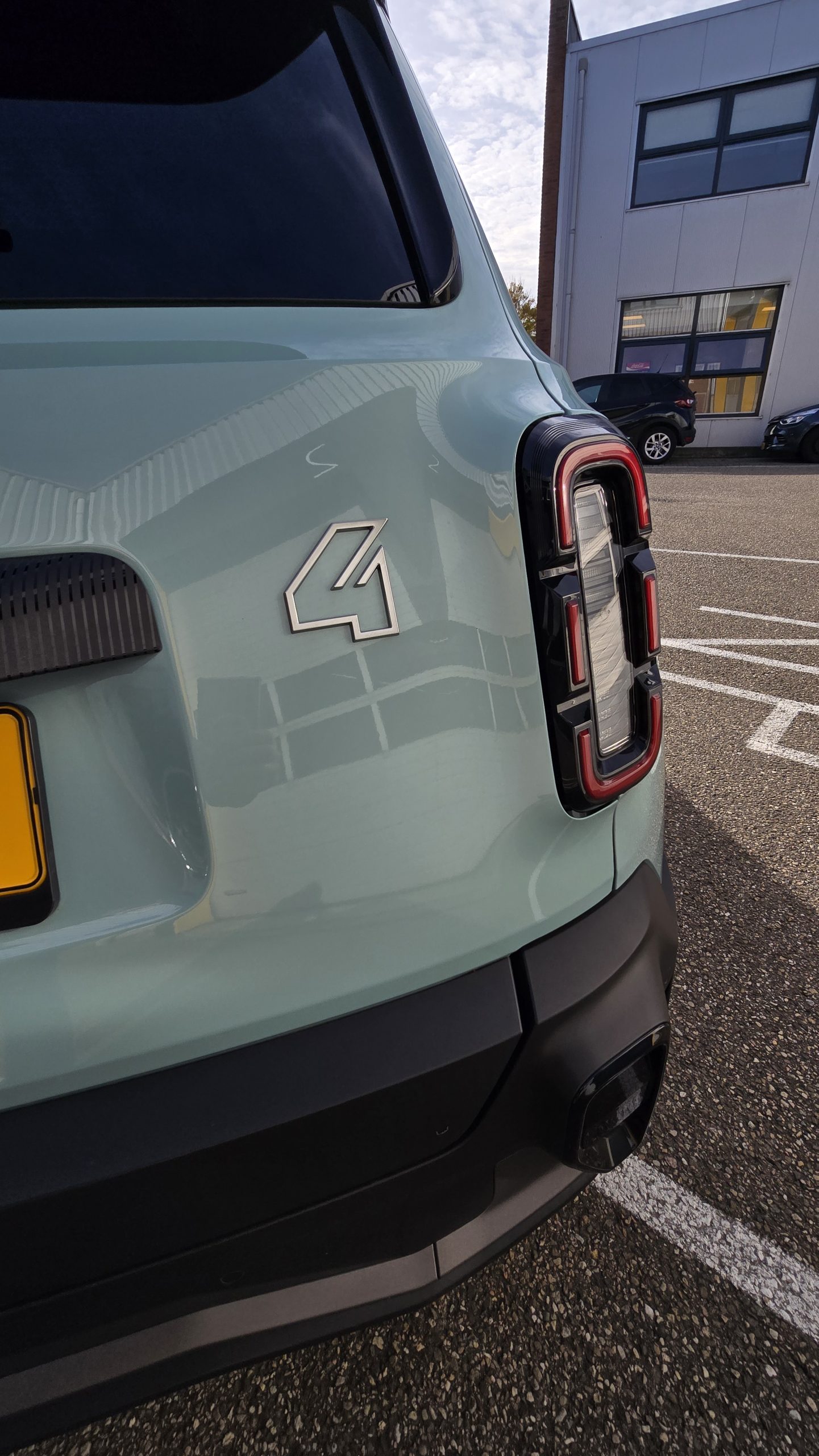 De achterkant van een lichtgroene elektrische auto met een moderne achterlamp en een bliksemschicht‑embleem, geparkeerd op een EV‑parkeerplaats bij een gebouw met grote ramen.