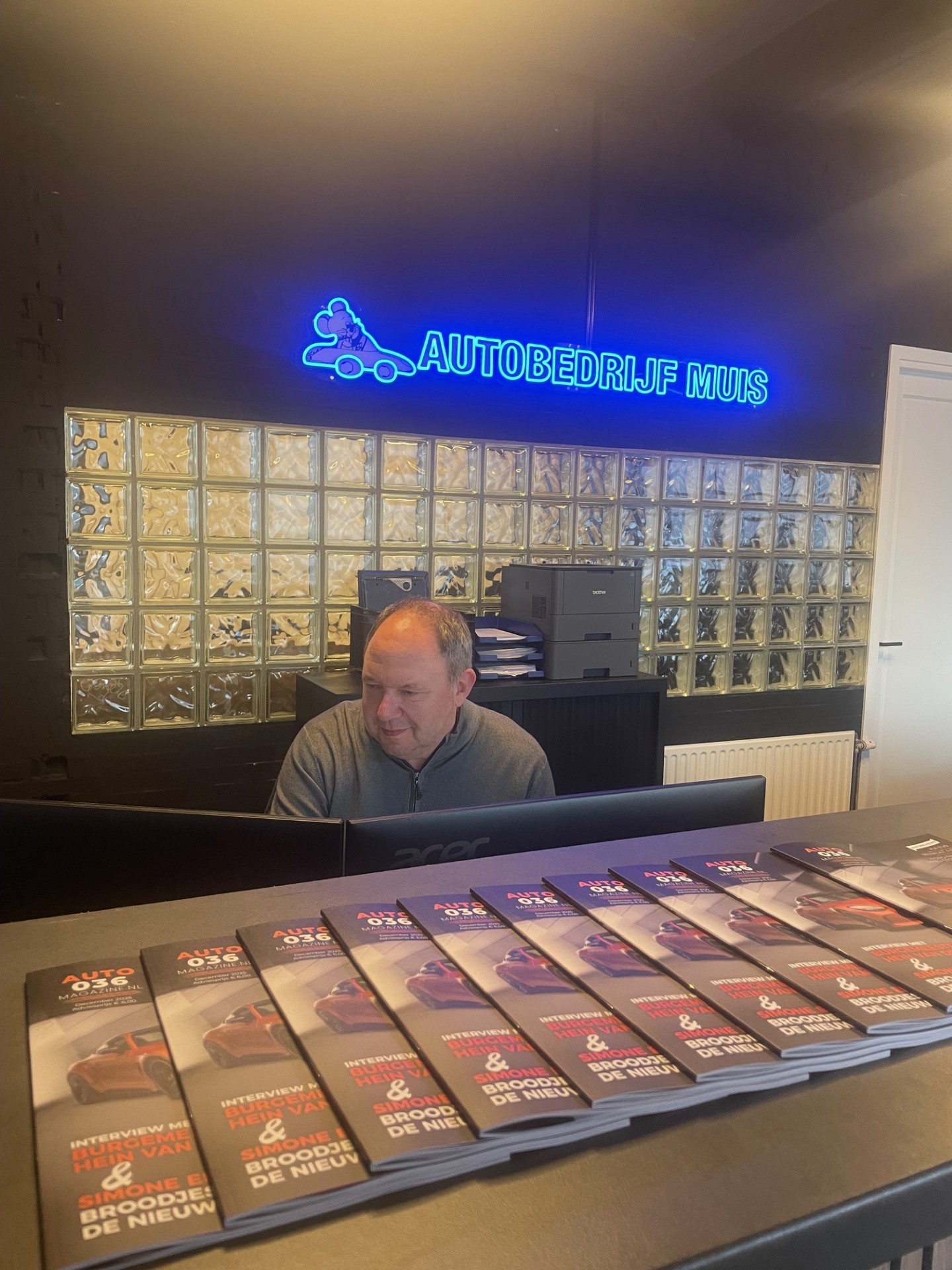 Man achter de balie van Autobedrijf Muis werkt aan een computer, met een neonlogo en stapels autobladen op de toonbank.