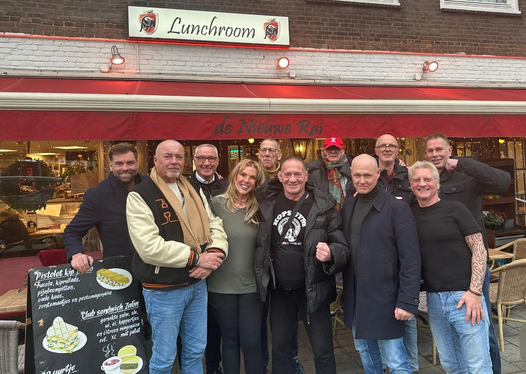 Een groep van twaalf mensen die poseert voor lunchroom De Nieuwe Rai, met een rood zonnescherm, winterkleding en een menubord met broodjes en koffieopties.