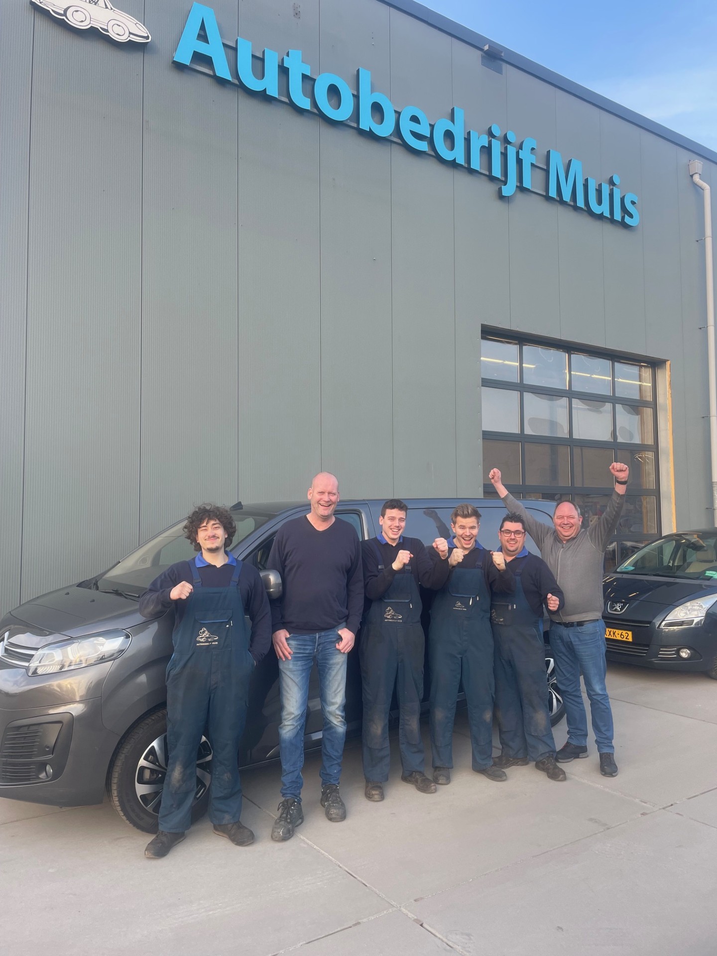 Groep medewerkers poseert lachend voor het gebouw van Autobedrijf Muis, met een busje en auto op de achtergrond.
