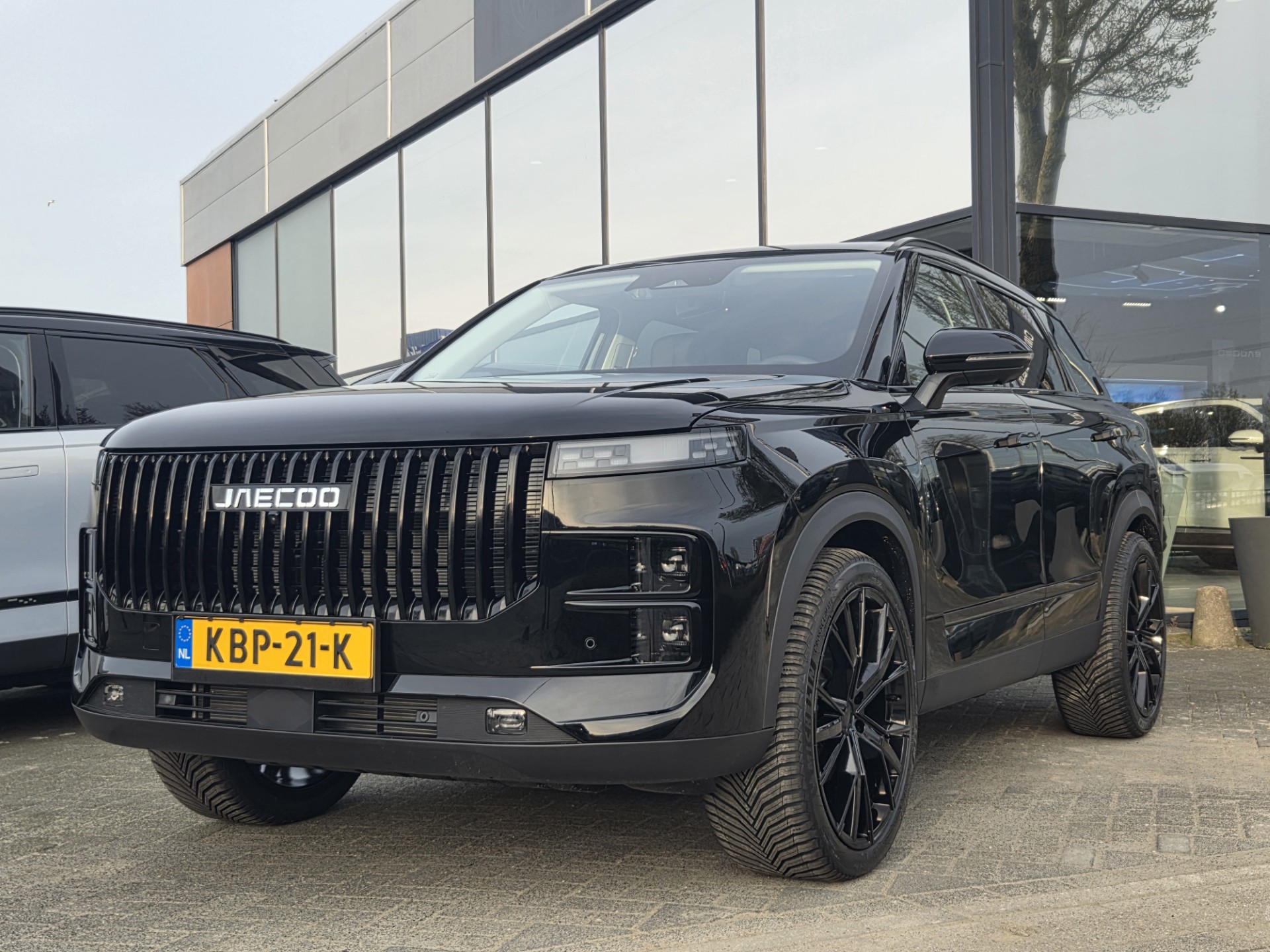 Een zwarte Jaecoo‑SUV geparkeerd voor een modern gebouw met grote glazen ramen, met een opvallende verticale grille en Nederlandse kentekenplaat.