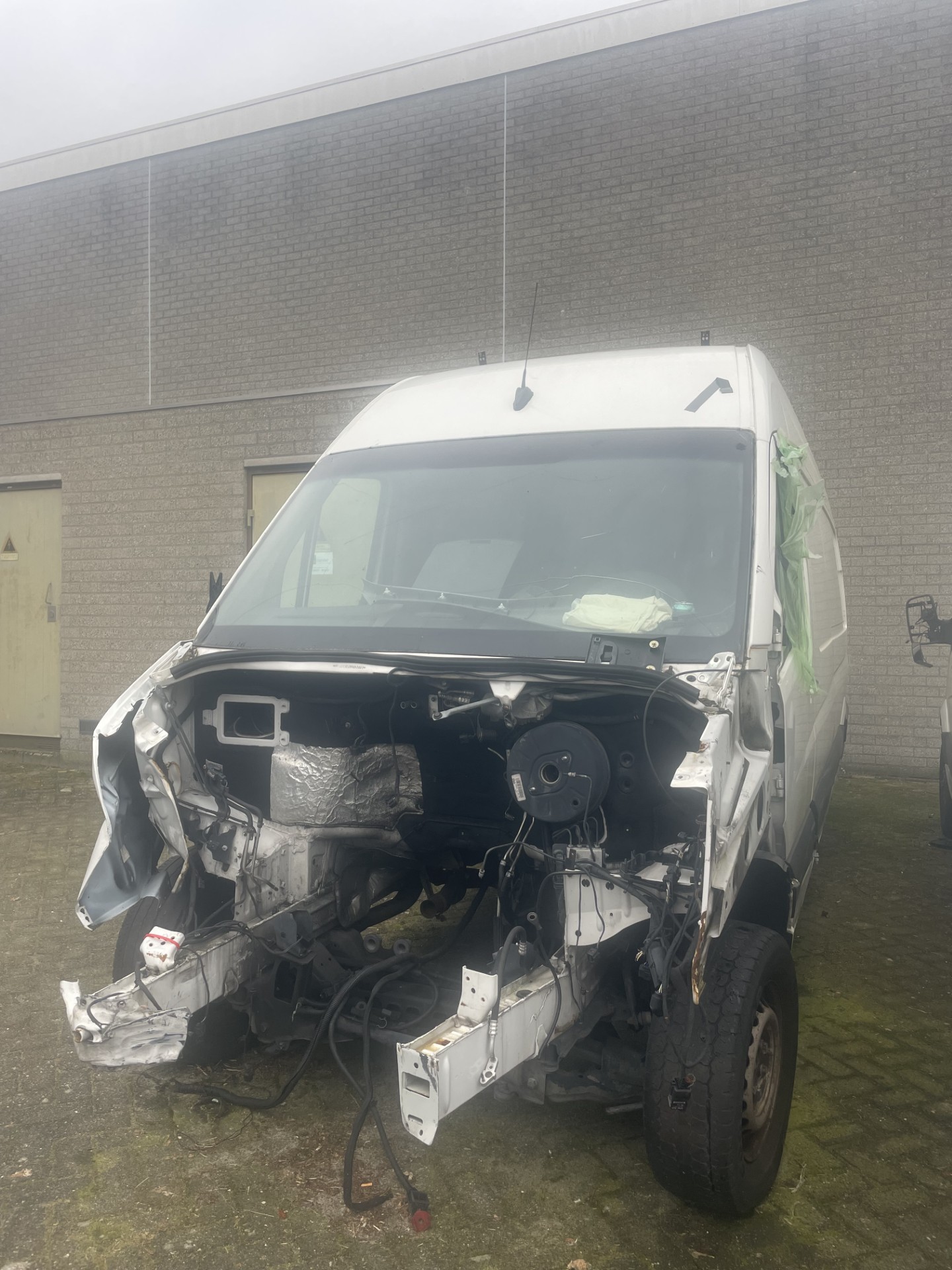 Een zwaar beschadigde witte bestelbus waarvan de volledige voorkant ontbreekt, met blootliggende onderdelen en een met plastic afgedekte bestuurdersruit