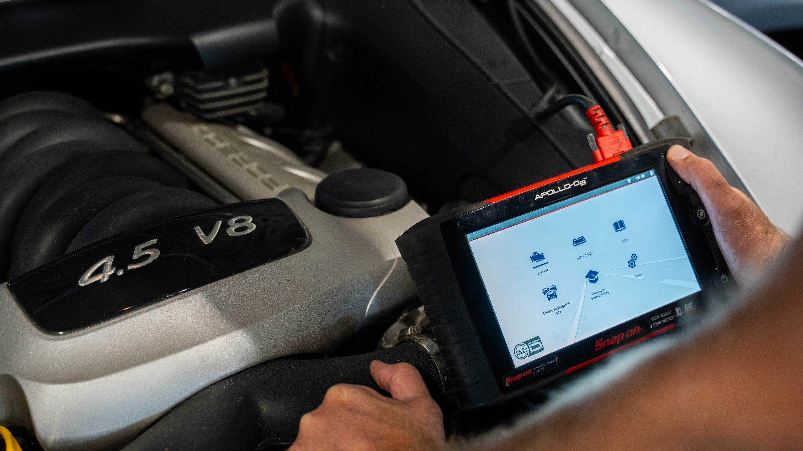 Monteur houdt een Snap-on APOLLO-D8 diagnosetablet vast boven de open motorkap van een auto met een 4.5 V8-motor.