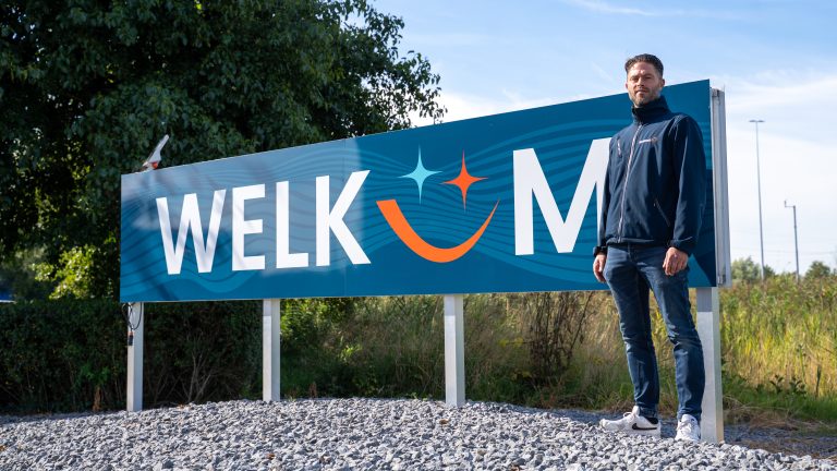 Man staat naast een groot welkomstbord met de tekst “WELKOM” en een glimlachend logo, geplaatst langs een weg met bomen en begroeiing op de achtergrond op een zonnige dag.