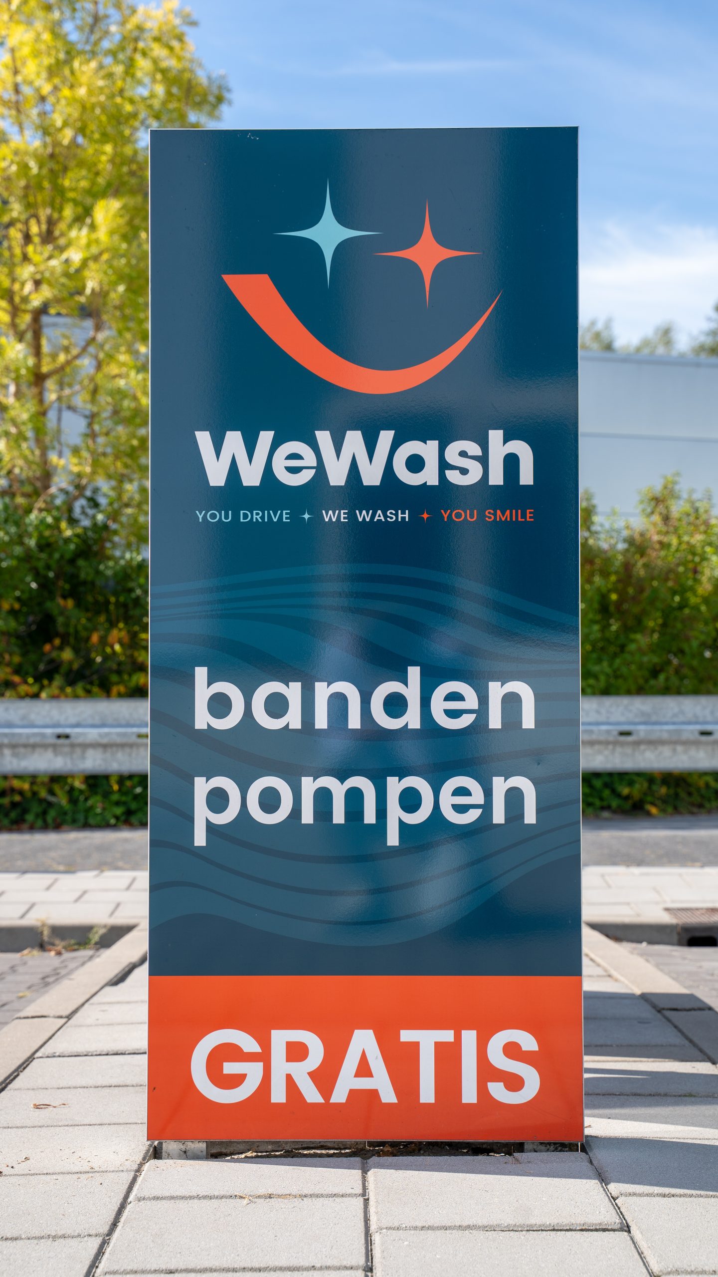Bord van WeWash met de tekst “banden pompen gratis”, geplaatst langs een parkeerplaats met bomen en groen op de achtergrond.