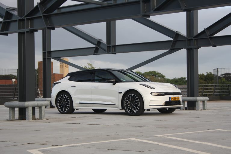 Een witte elektrische auto met zwarte accenten staat geparkeerd in een open parkeergarage, met metalen constructiebalken en een deels bewolkte lucht op de achtergrond.