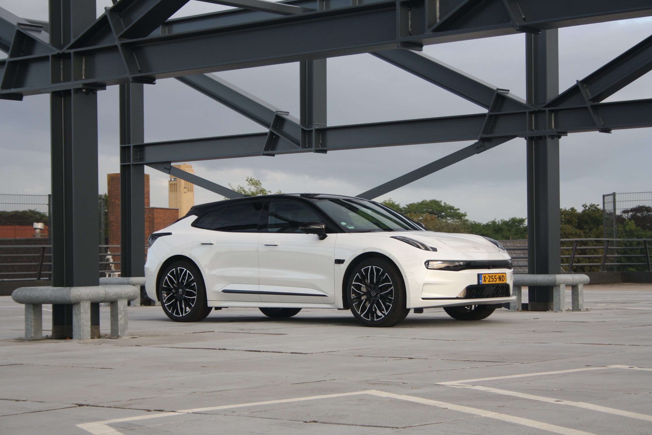 Een witte elektrische auto met zwarte accenten staat geparkeerd in een open parkeergarage, met metalen constructiebalken en een deels bewolkte lucht op de achtergrond.