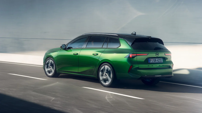 Een groene Opel Astra Sports Tourer die rijdend over een meerbaansweg is vastgelegd, met een zwarte daklijn en een vervaagde stedelijke achtergrond.