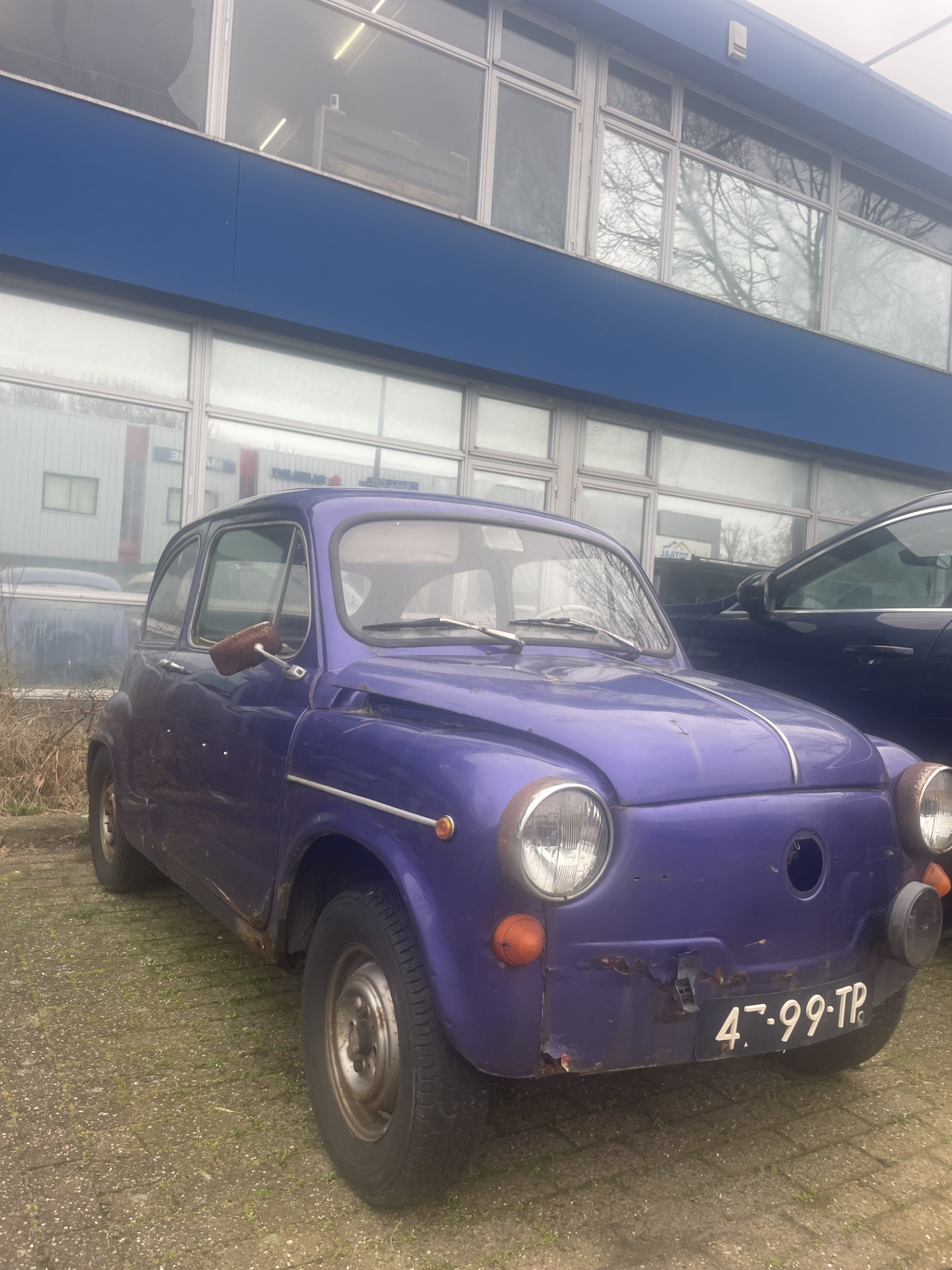Een paarse vintage auto met roestplekken, geparkeerd op een stenen plein voor een modern gebouw met grote ramen.