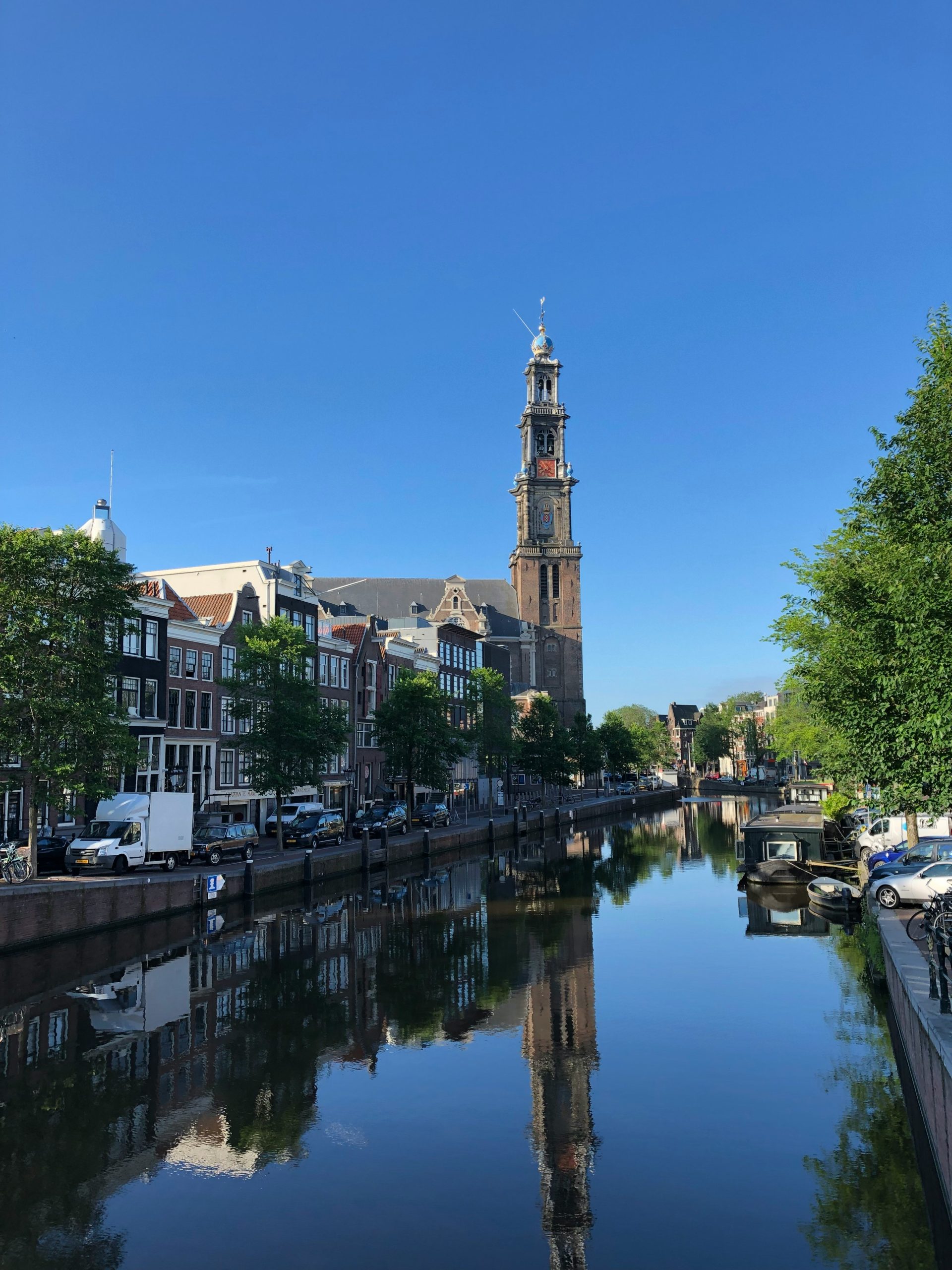Een rustige Amsterdamse gracht met de Westerkerk die weerspiegelt in het water, omringd door grachtenpanden, bomen, geparkeerde auto’s en boten onder een heldere blauwe lucht.