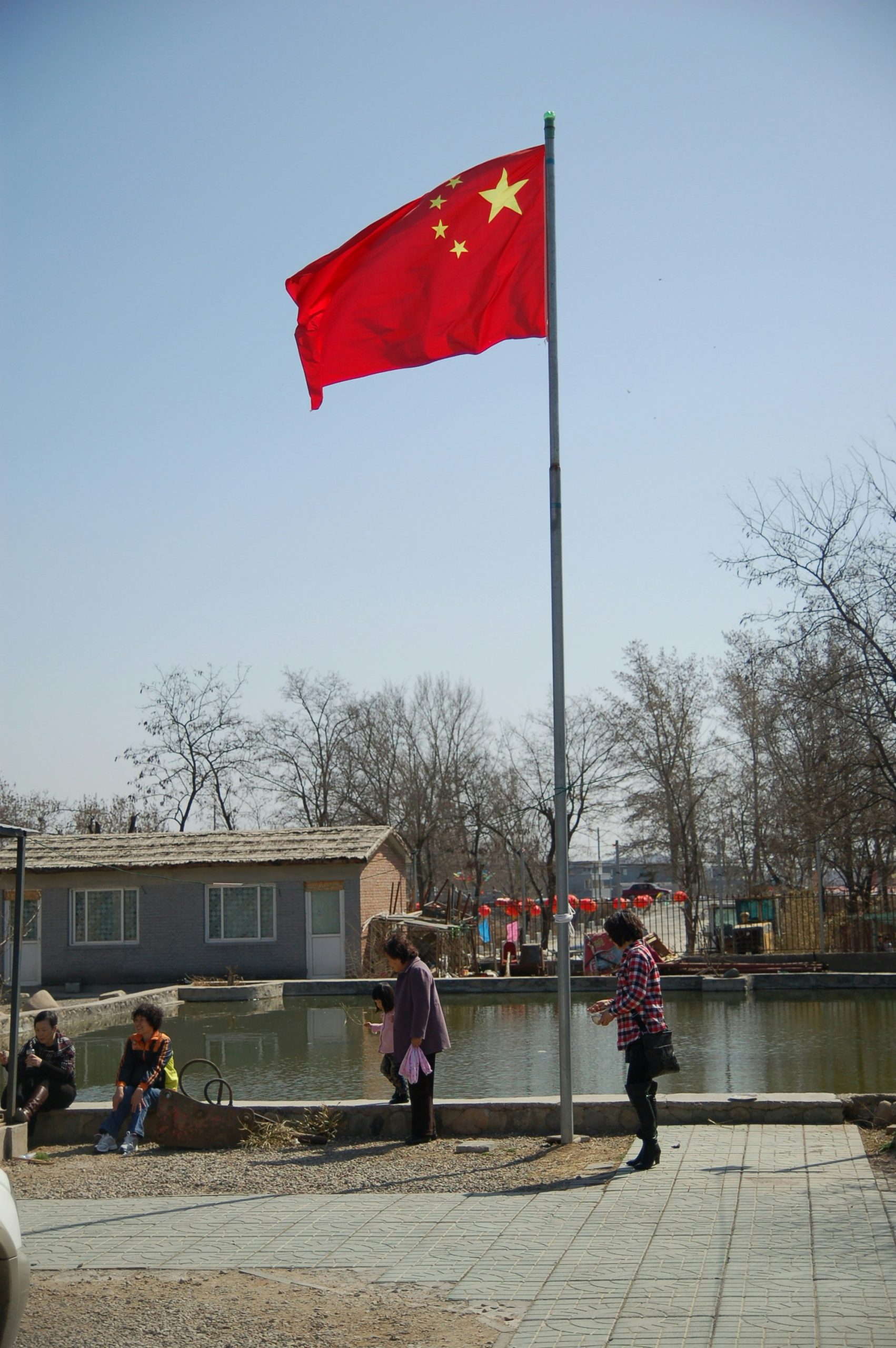 Een wapperende Chinese vlag aan een hoge vlaggenmast bij een vijver, met mensen op de oever, een gebouw met pannendak en rode lantaarns op de achtergrond.