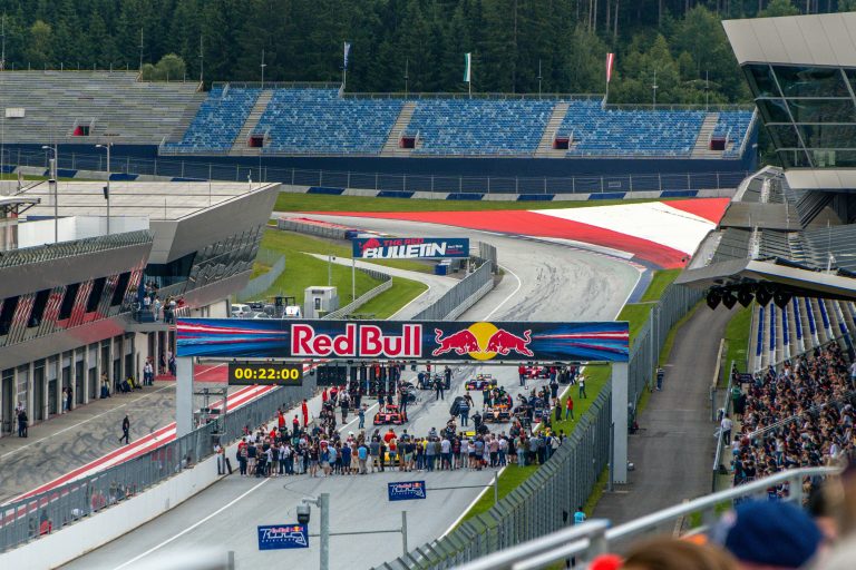 Raceauto’s staan opgesteld op de startgrid van een circuit, omringd door teams en toeschouwers.