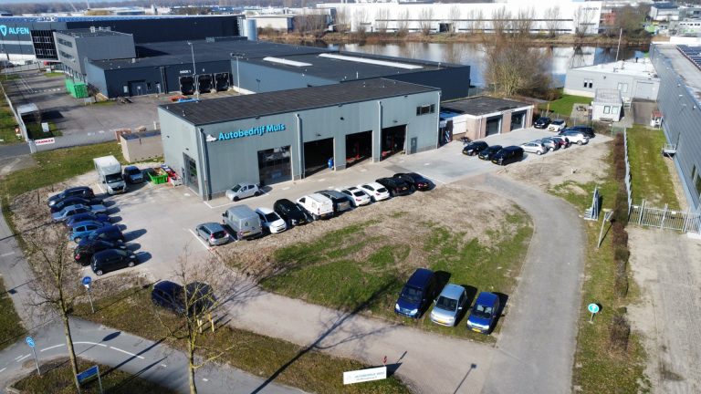 Luchtfoto van een bedrijventerrein met Autobedrijf Muis en het naastgelegen Alfen‑gebouw, omringd door geparkeerde auto’s en wegen.