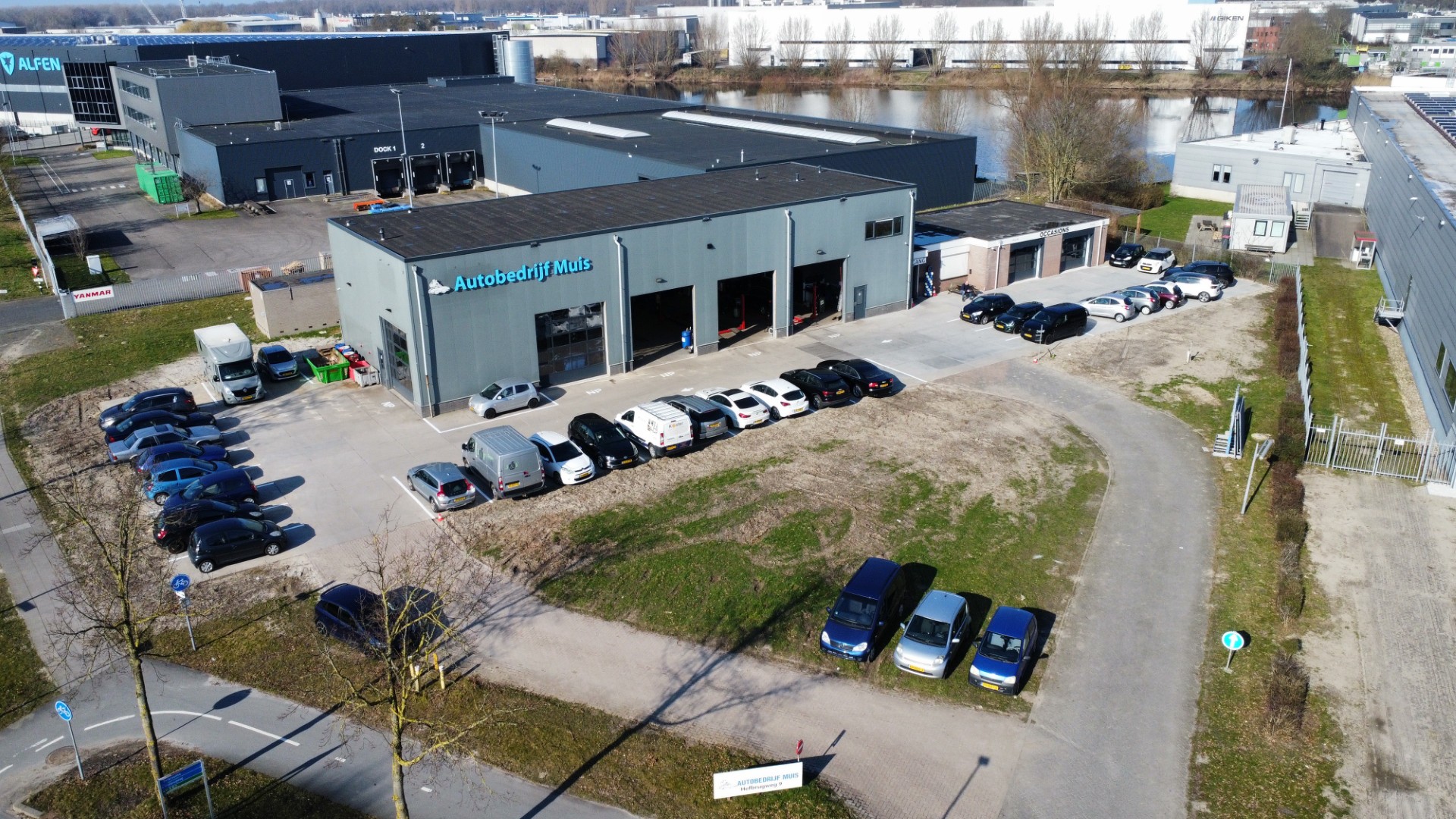 Luchtfoto van een bedrijventerrein met Autobedrijf Muis en het naastgelegen Alfen‑gebouw, omringd door geparkeerde auto’s en wegen.