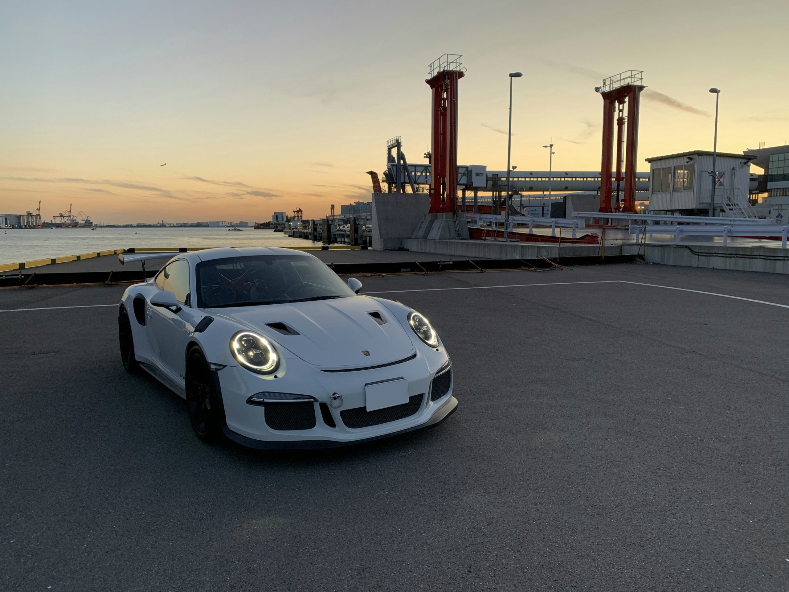 Een witte Porsche 911 GT3 RS geparkeerd bij een havengebied tijdens zonsondergang, met industriële kranen op de achtergrond en de koplampen aan.