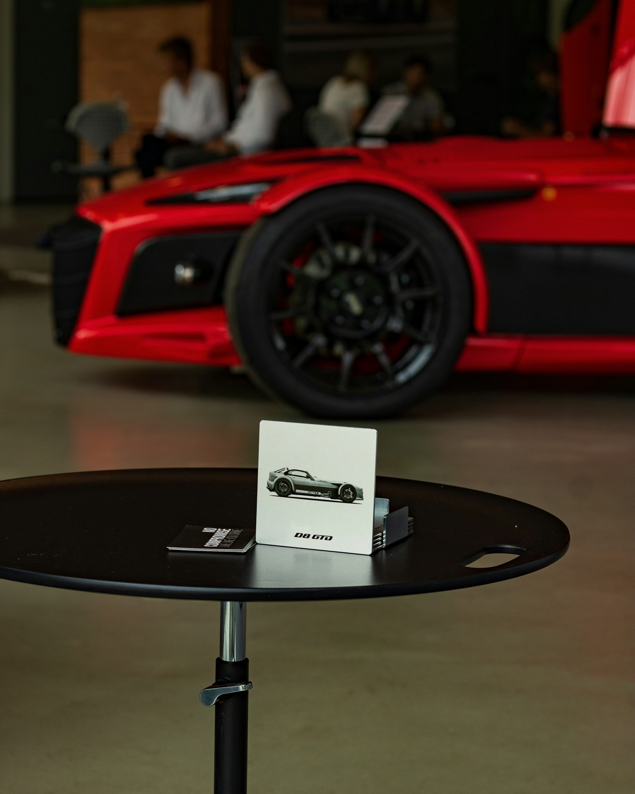 Een showroomtafel met een displaykaart van een sportwagen en op de achtergrond een felrode sportauto met zwarte velgen en mensen die zitten en praten.