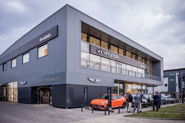 Moderne Broekhuis‑dealer met Hyundai‑ en Genesis‑signing, glazen gevel en bezoekers bij geparkeerde auto's.