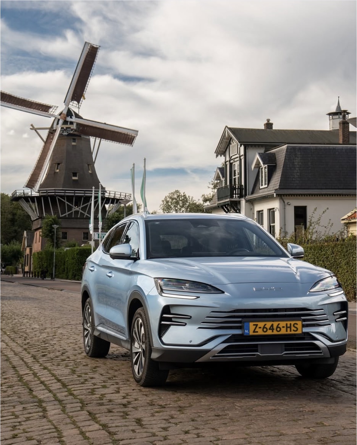 Moderne lichtblauwe SUV geparkeerd op een kasseienstraat voor een traditionele Nederlandse molen en huizen.