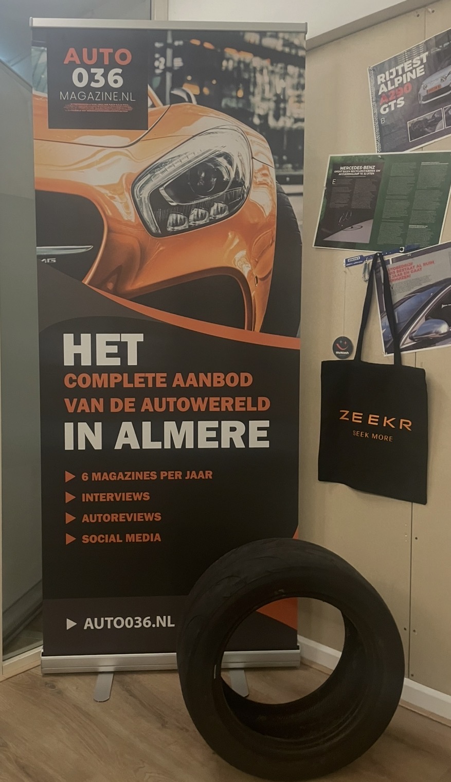Promotiewand van Auto036 Magazine met close‑up van een oranje sportauto, omringd door automotive posters en een band op de vloer.