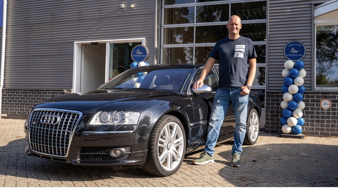 Persoon naast een zwarte Audi S8 voor een moderne dealergevel, met blauwe‑witte ballonnen en een welkomstbord.