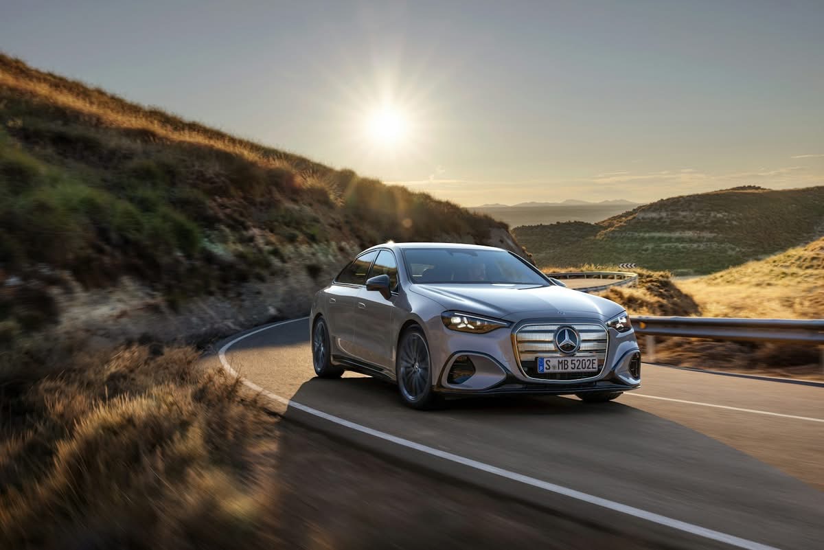Zilveren Mercedes‑Benz sedan rijdend over een bochtige bergweg in warm avondlicht met heuvelachtig landschap.