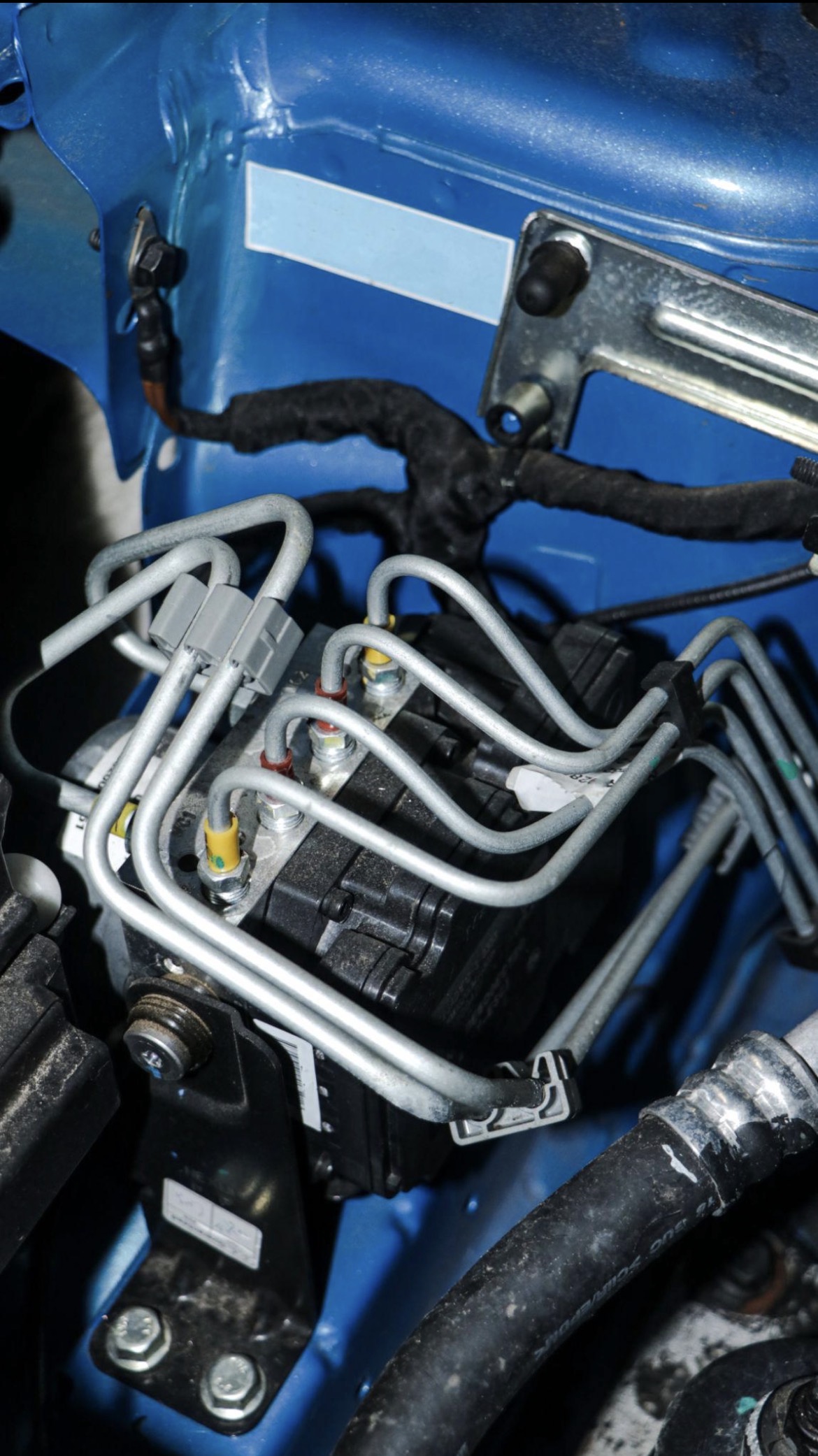 Close‑up van een ABS‑module in de motorruimte met metalen remleidingen, stekkers en blauwe carrosseriedelen.