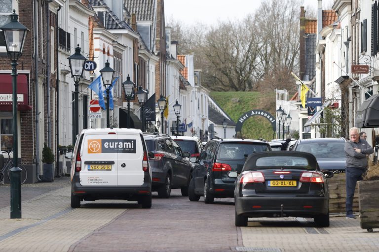 Smalle Europese straat met geparkeerde auto's, waaronder een Eurami‑bestelbus, omringd door winkels en historische gevels.