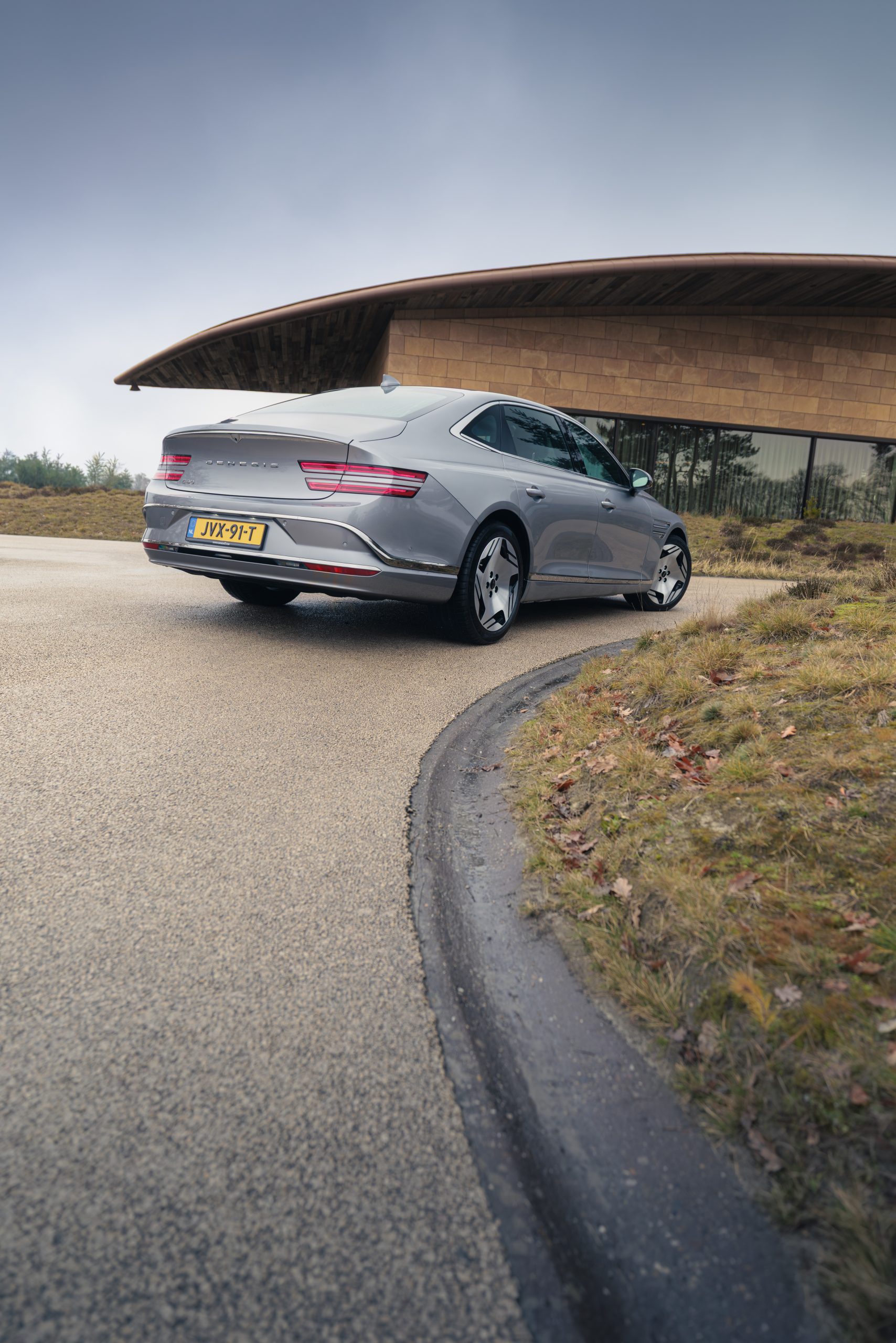 Luxe elektrische sedan, Genesis G80 Electrified, geparkeerd op een gebogen weg bij een modern gebouw met een opvallend houten dak, gezien van achteren.