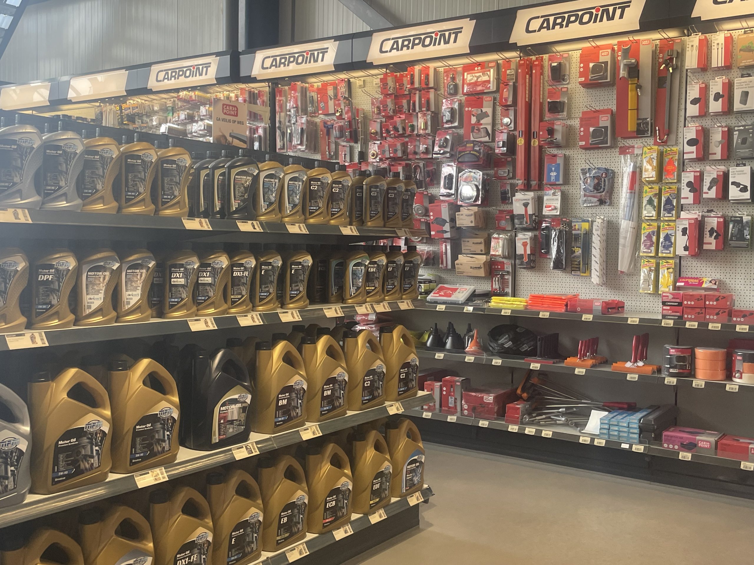 Schappen in een automaterialenzaak met motorolie in goud‑zwarte flessen en Carpoint‑accessoires in rood‑witte verpakkingen