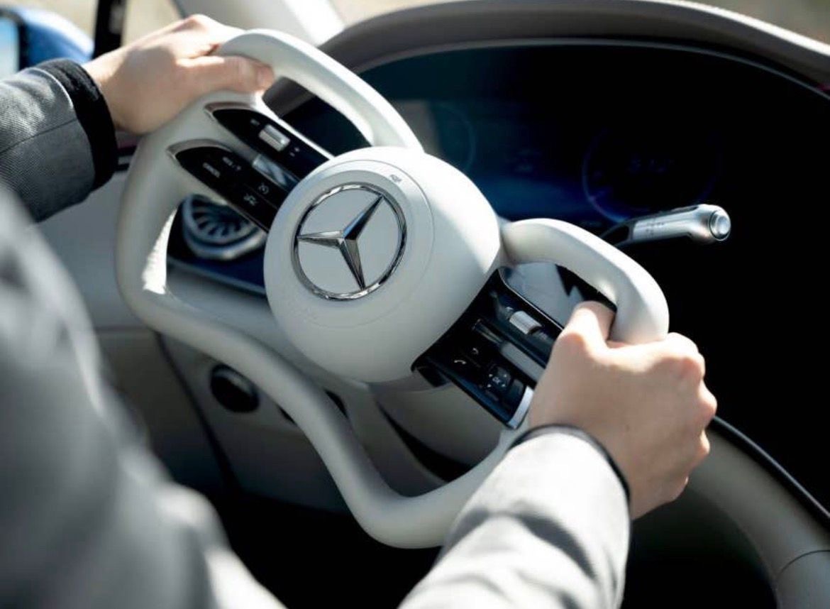 Een close‑up van handen die een futuristisch, rechthoekig stuurwiel met Mercedes‑logo vasthouden in een moderne auto‑interieur.