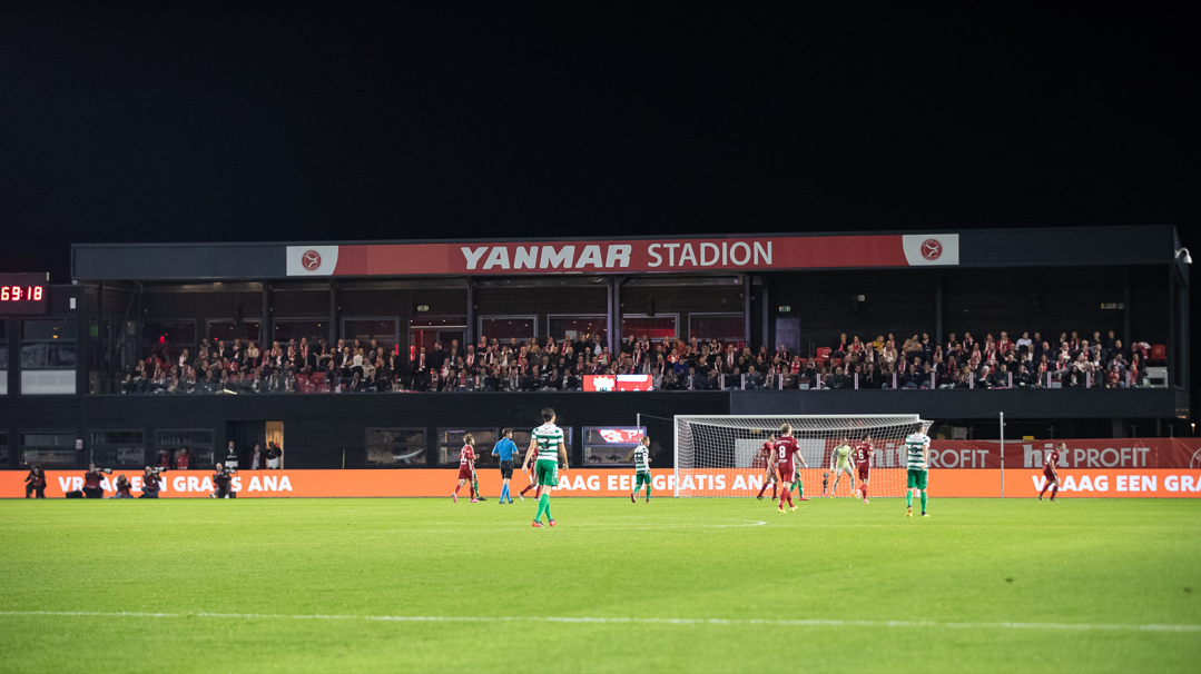 Avondwedstrijd in het Yanmar Stadion met spelers op het veld, volle tribunes en verlichte reclameborden langs de zijlijn.