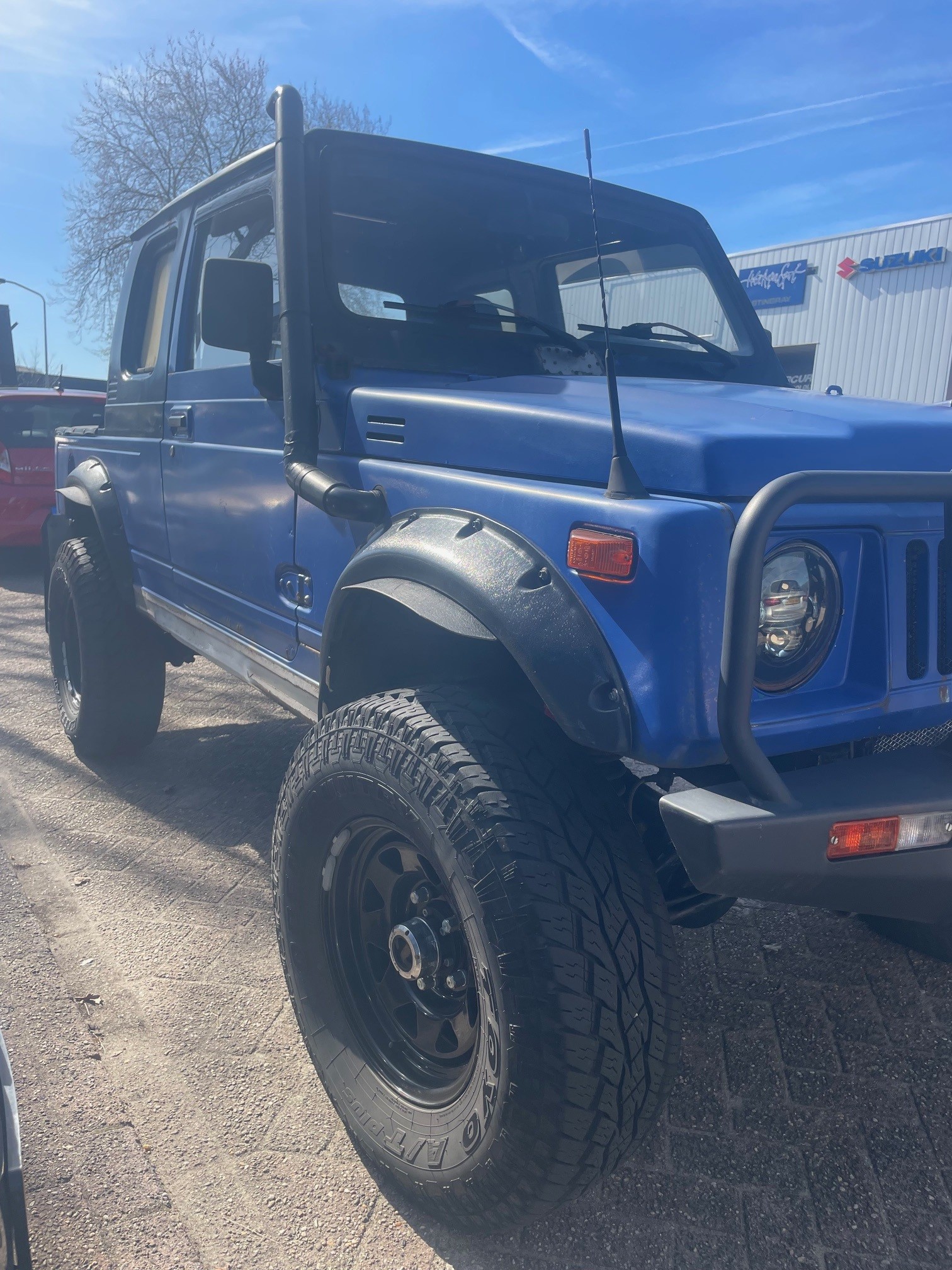 Een blauwe, aangepaste Suzuki‑terreinwagen met grote off‑roadbanden, snorkel en bullbar, geparkeerd buiten.