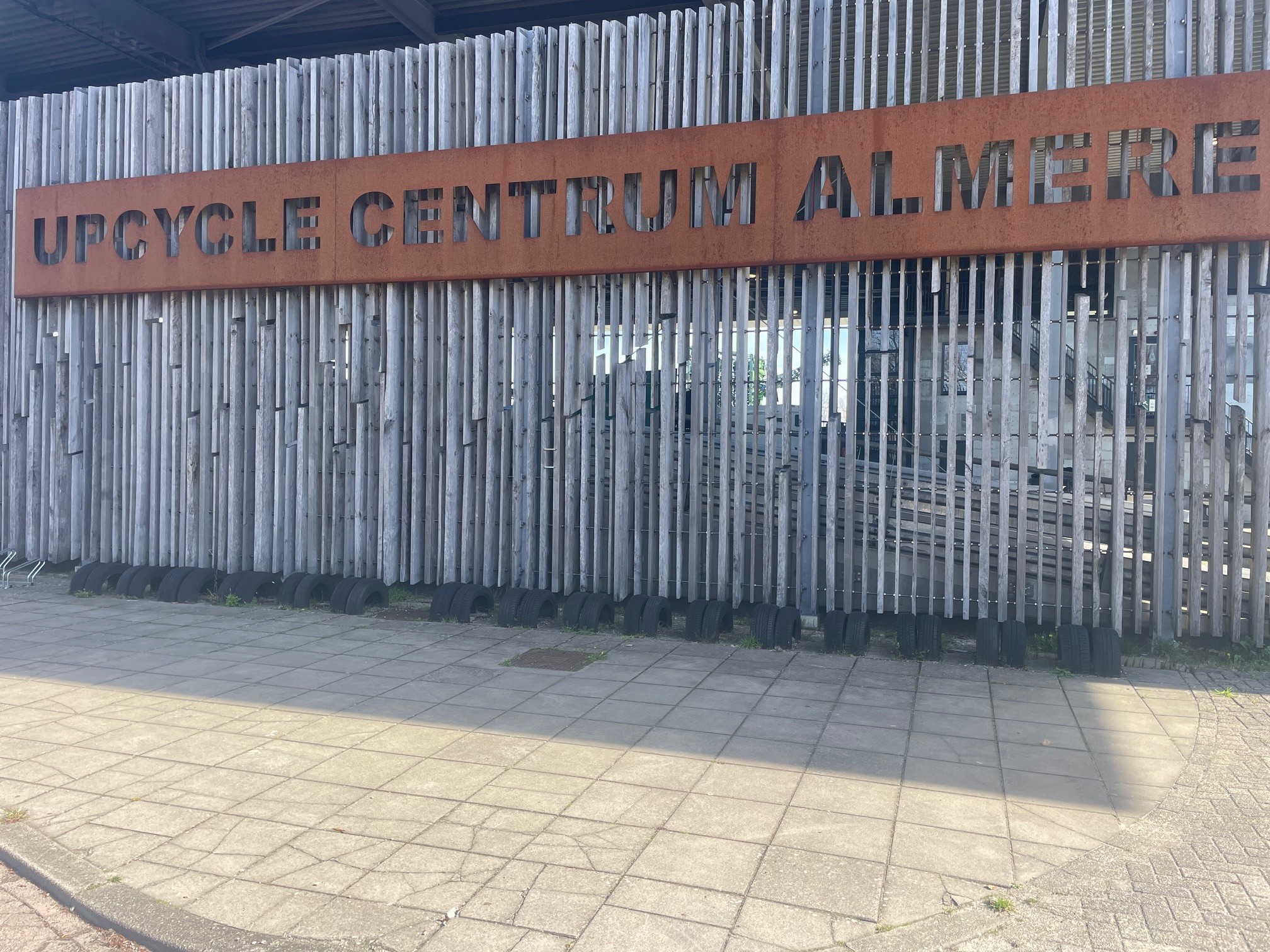 De houten gevel van het Upcycle Centrum Almere met een roestkleurig bord waarop ‘Upcycle Centrum Almere’ staat.