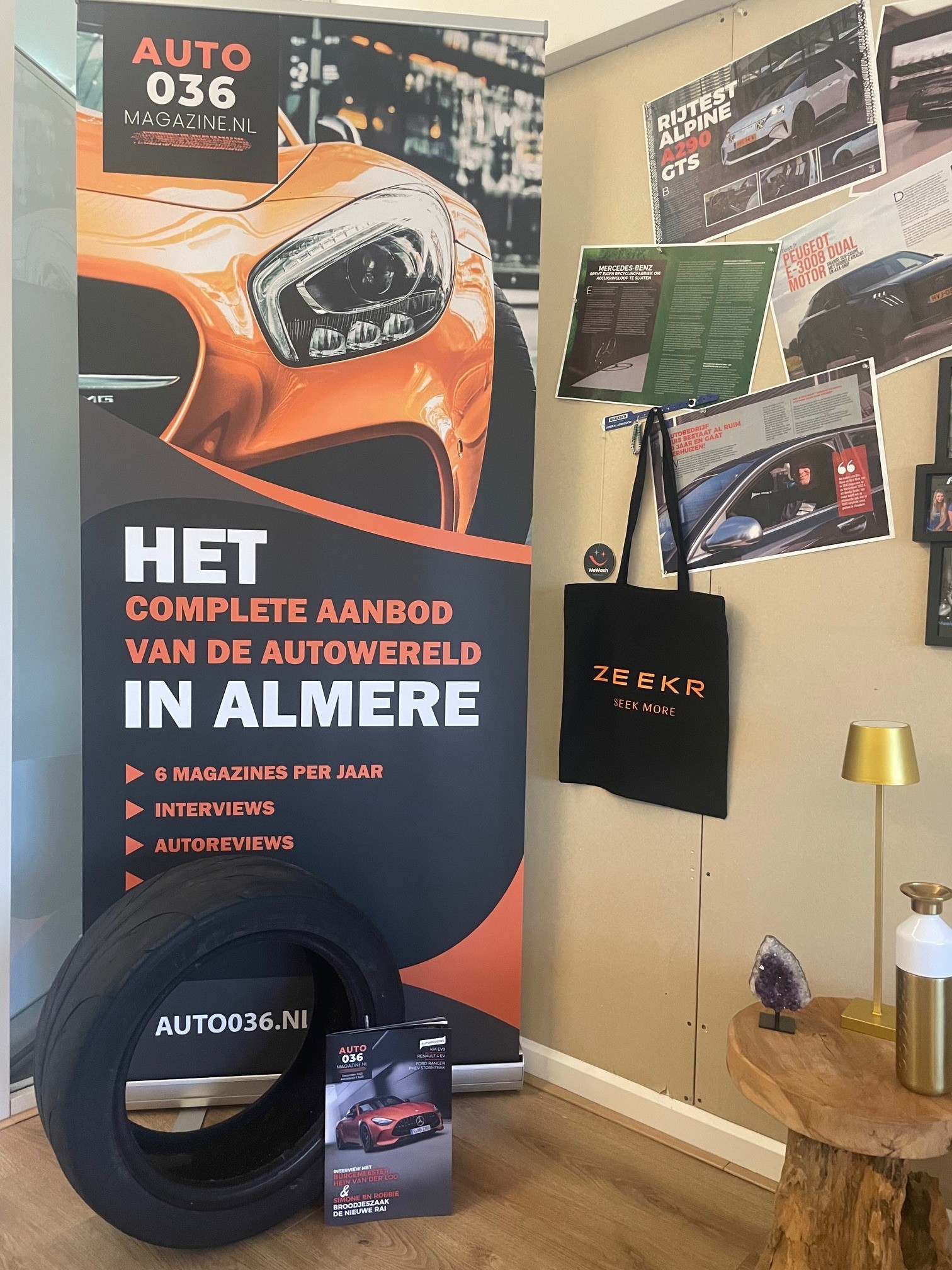Een kleine indoor display met een grote banner van AUTO036 Magazine, een autoband, een tijdschriftenstandaard en autogerelateerde posters aan de muur.