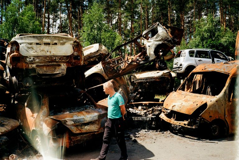 Persoon die langs een grote stapel verbrande en verroeste autowrakken loopt, omringd door bomen en groen.