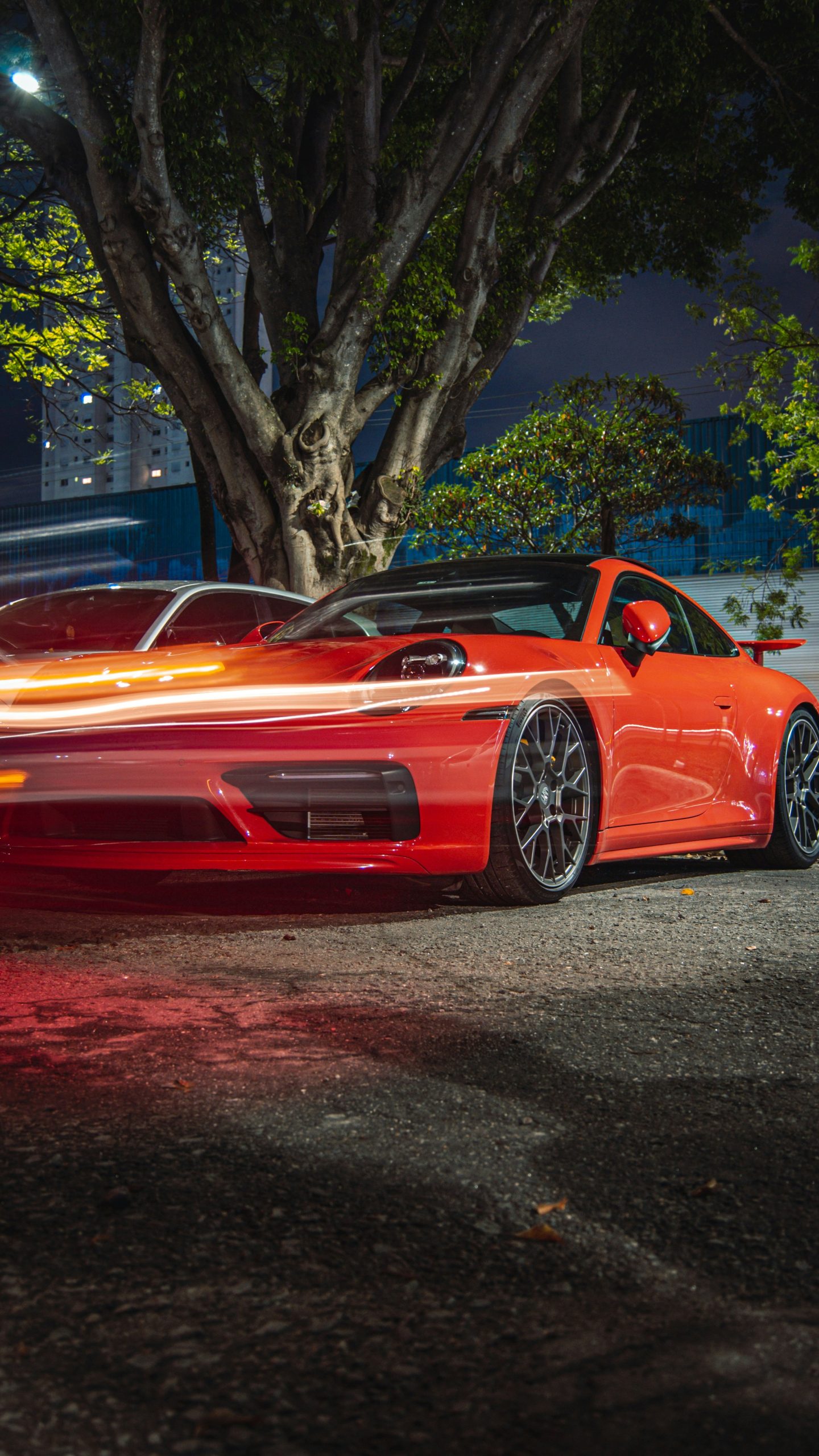 Rode sportauto geparkeerd op straat in de avond, met lichtstrepen van passerend verkeer en bomen op de achtergrond.