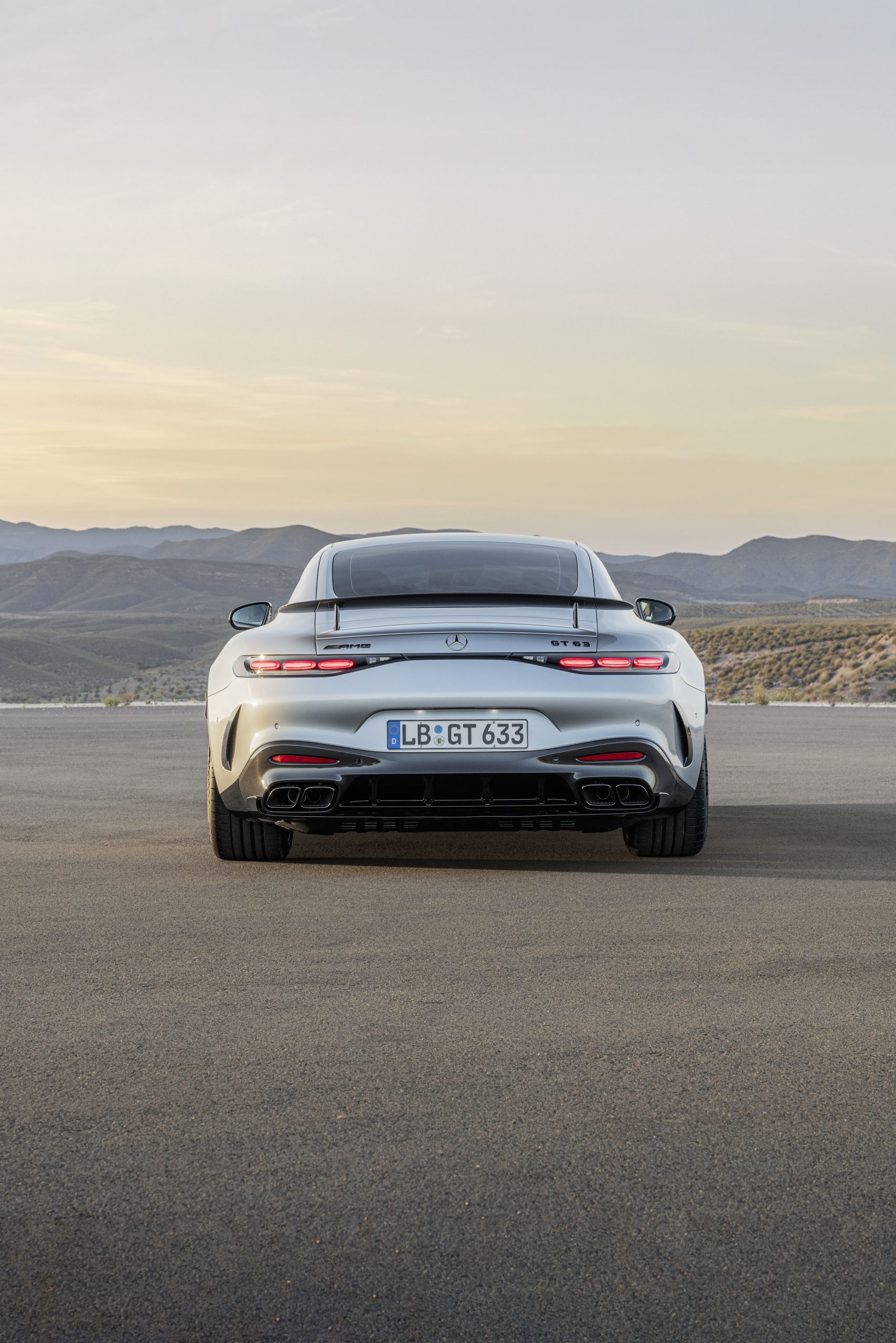 Achterkant van een zilveren sportauto (Mercedes-Benz AMG GT) op een lege weg in een bergachtig landschap bij zonsondergang.