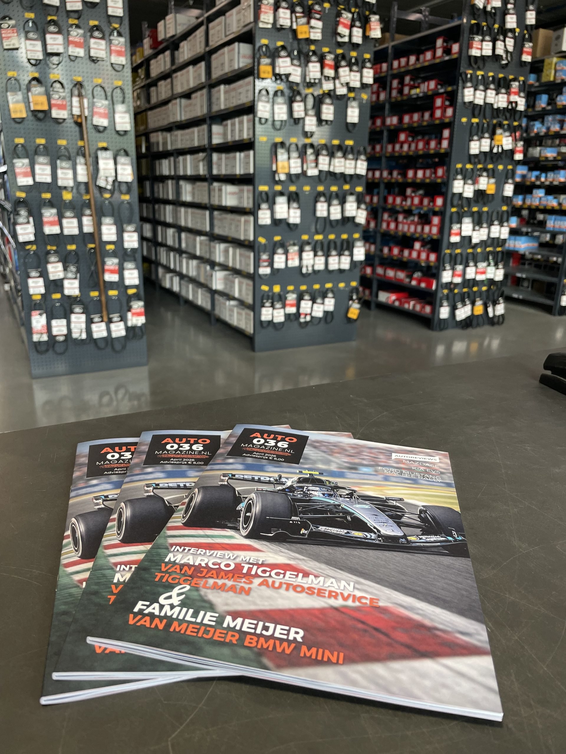 Magazines met Formule‑1 cover op een balie in een automaterialenzaak, met op de achtergrond schappen vol onderdelen