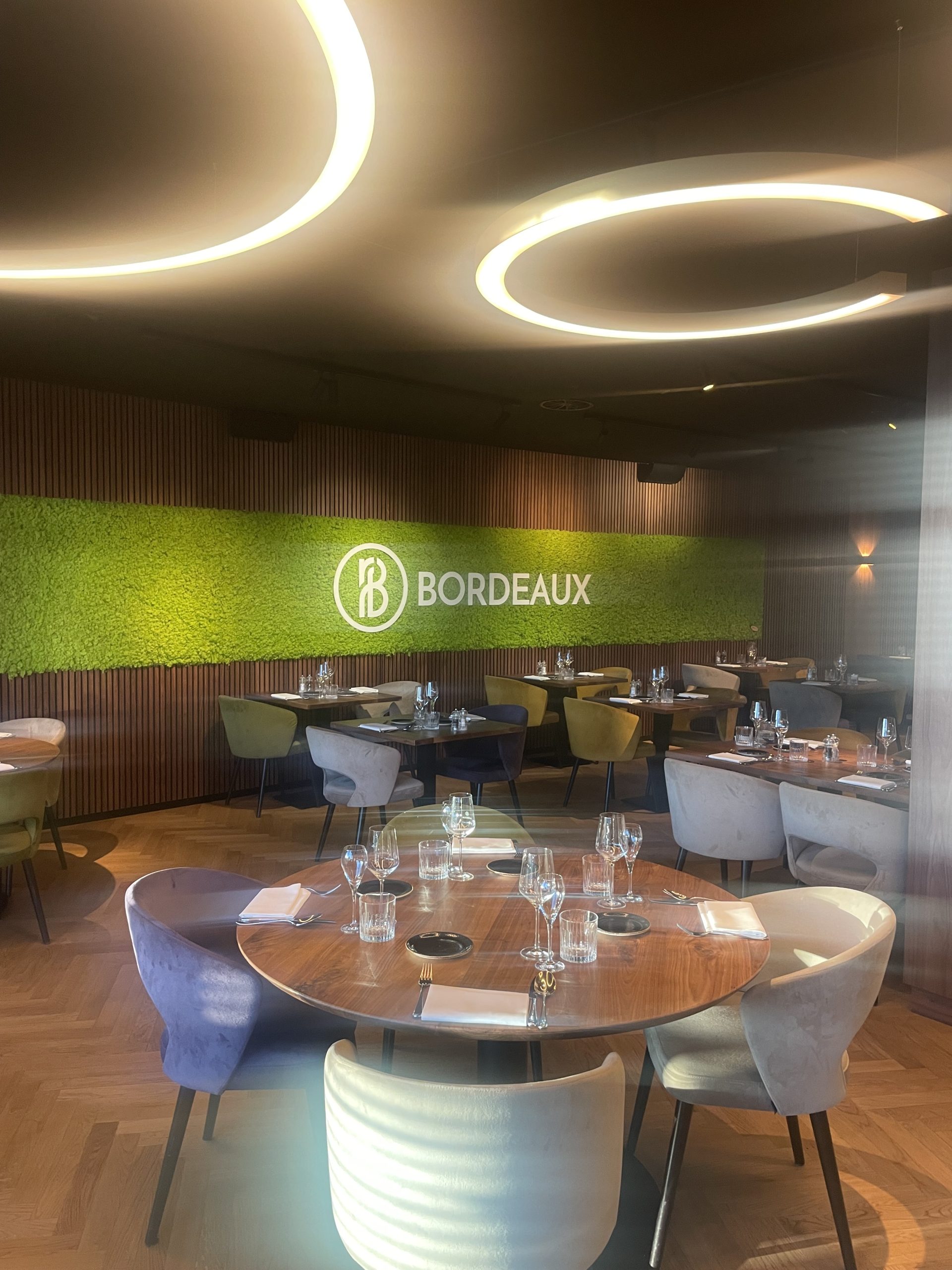 Stijlvol restaurantinterieur met ronde gedekte tafels, kleurrijke stoelen en groene wand met Bordeaux‑logo.