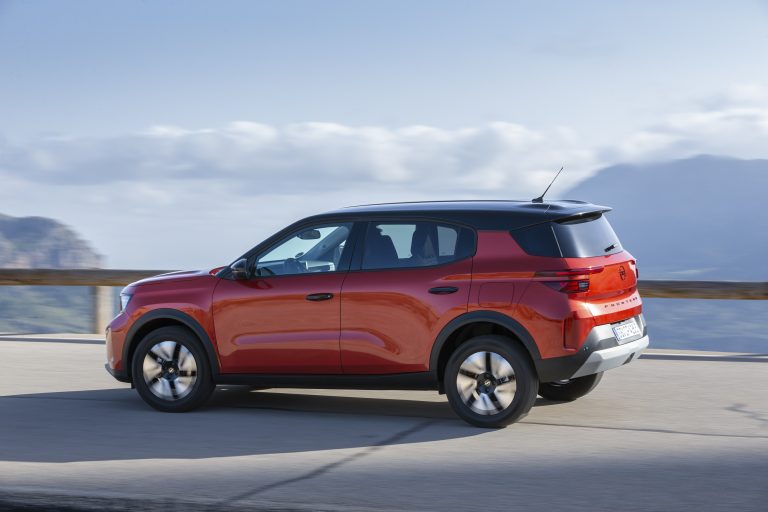 Rode compacte SUV rijdend over een bergweg met zwarte daklijn en heuvelachtig landschap op de achtergrond.