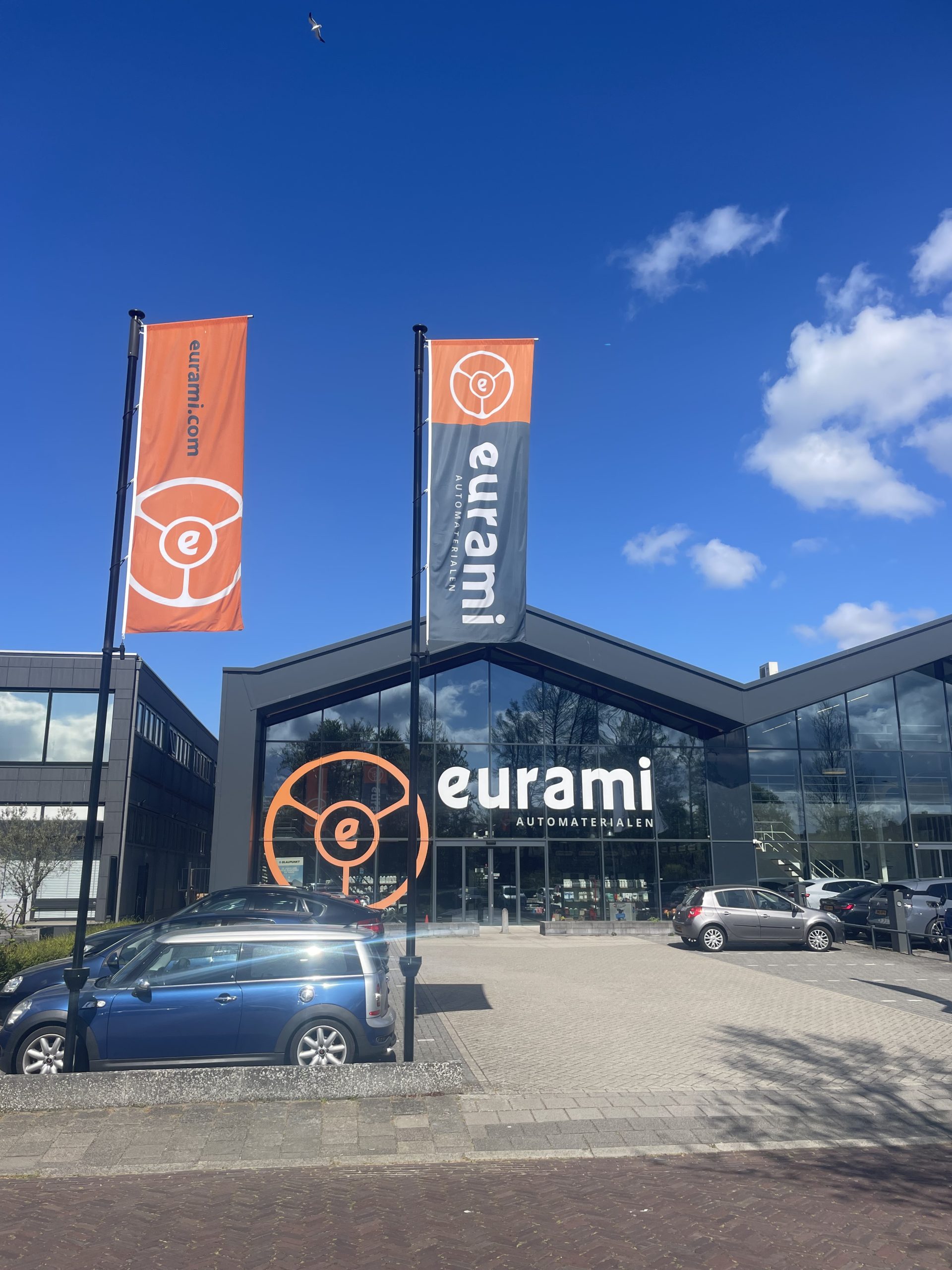 Modern pand met grote glasgevel en Eurami‑branding, met vlaggen en geparkeerde auto's voor de ingang.