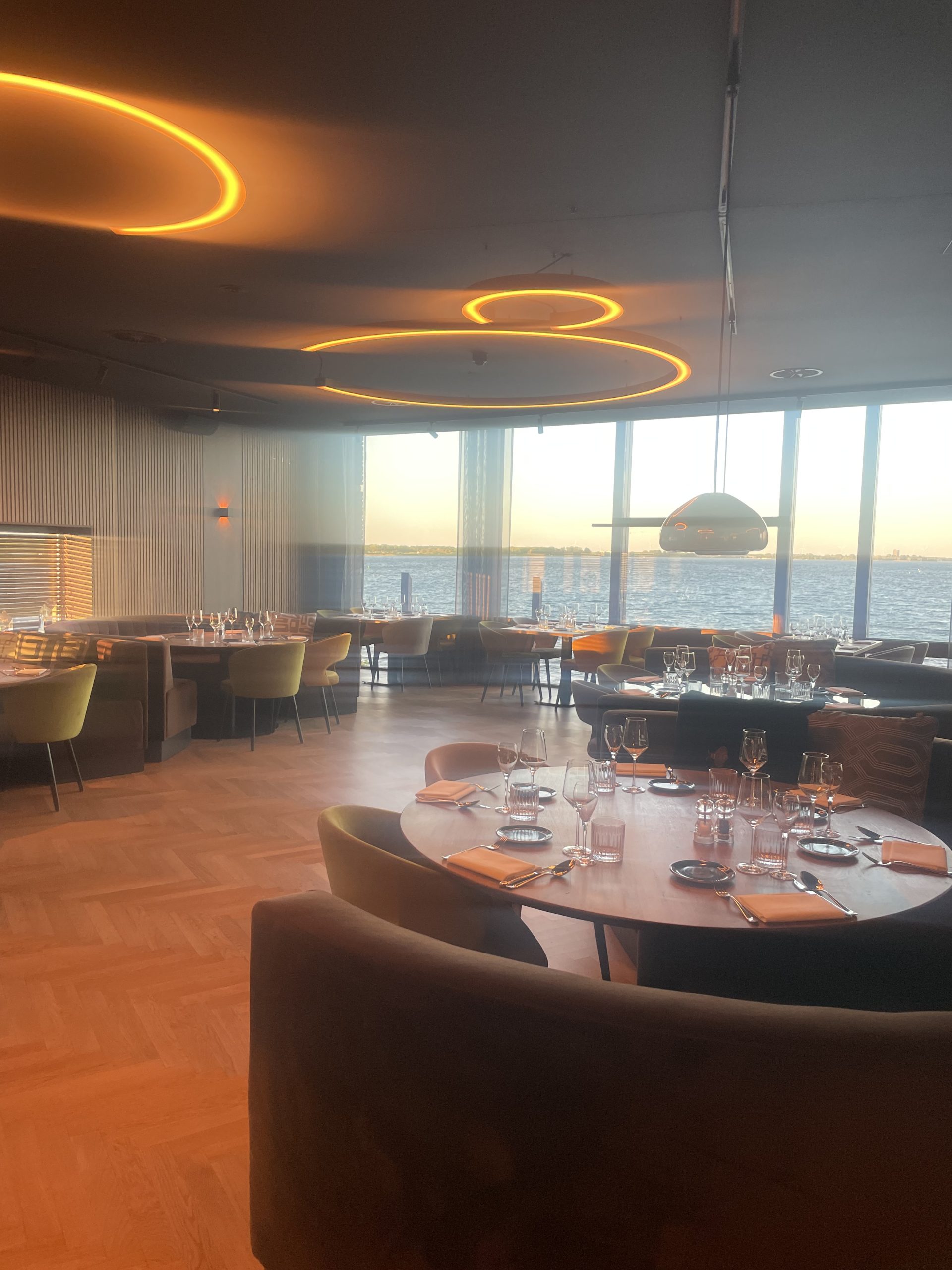 Modern restaurant met ronde houten tafels, warme verlichting en panoramisch uitzicht op water en skyline bij zonsondergang.