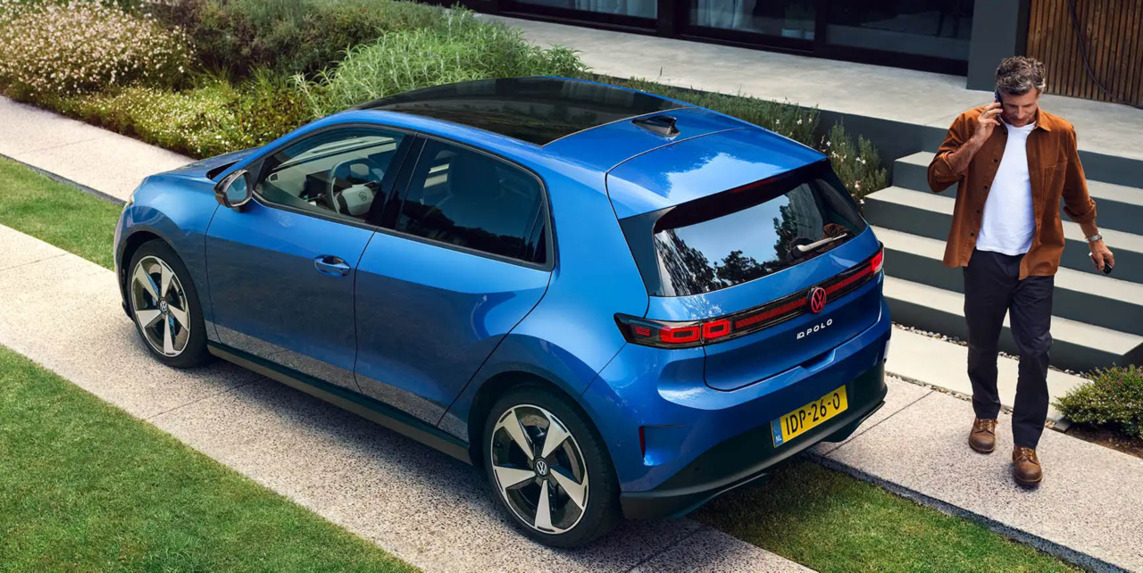 Blauwe Volkswagen Polo met zwart dak geparkeerd naast een moderne woning, terwijl een persoon erlangs loopt.