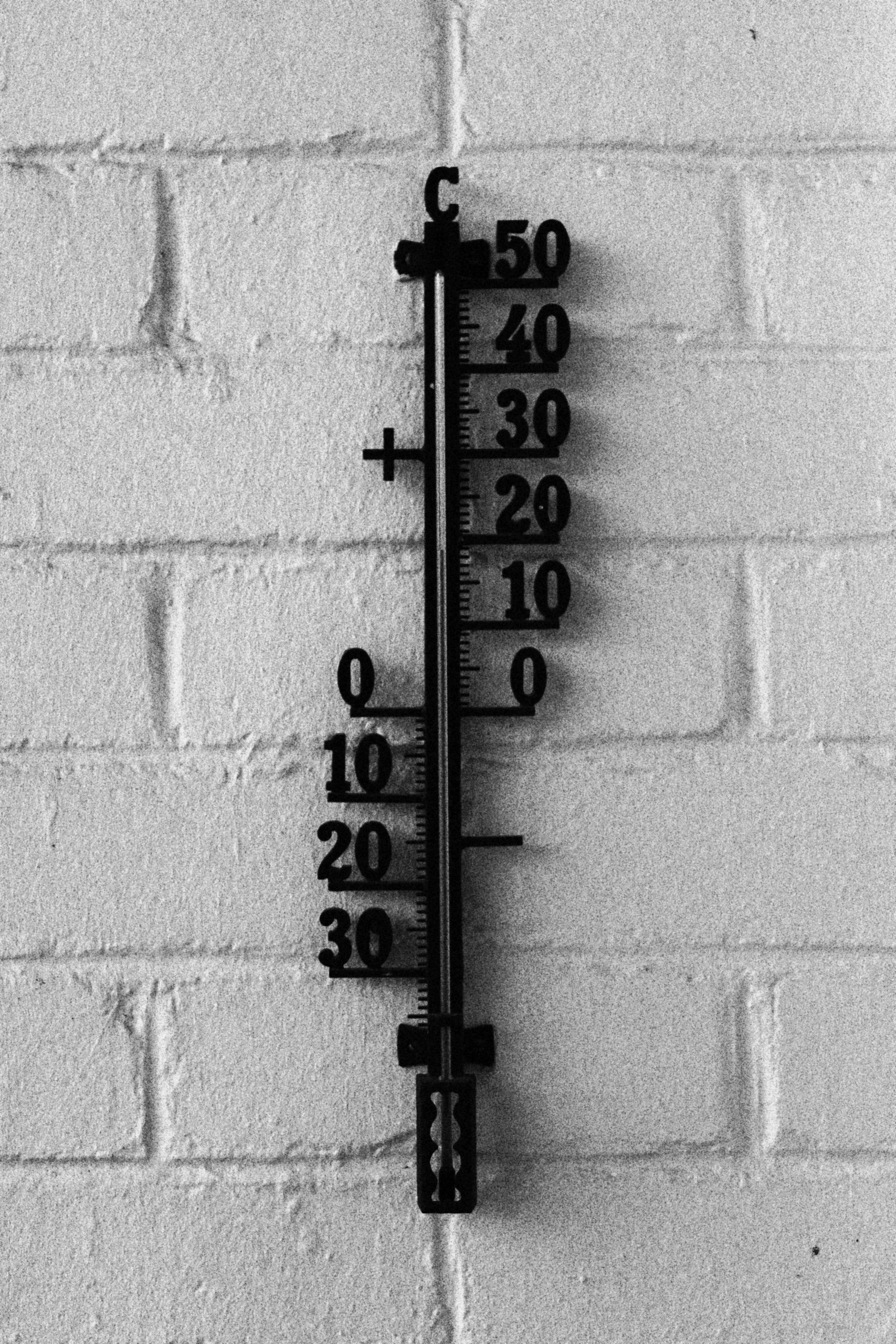 Zwarte metalen buitenthermometer tegen een lichte bakstenen muur met schaal van −30 tot +50 graden Celsius.