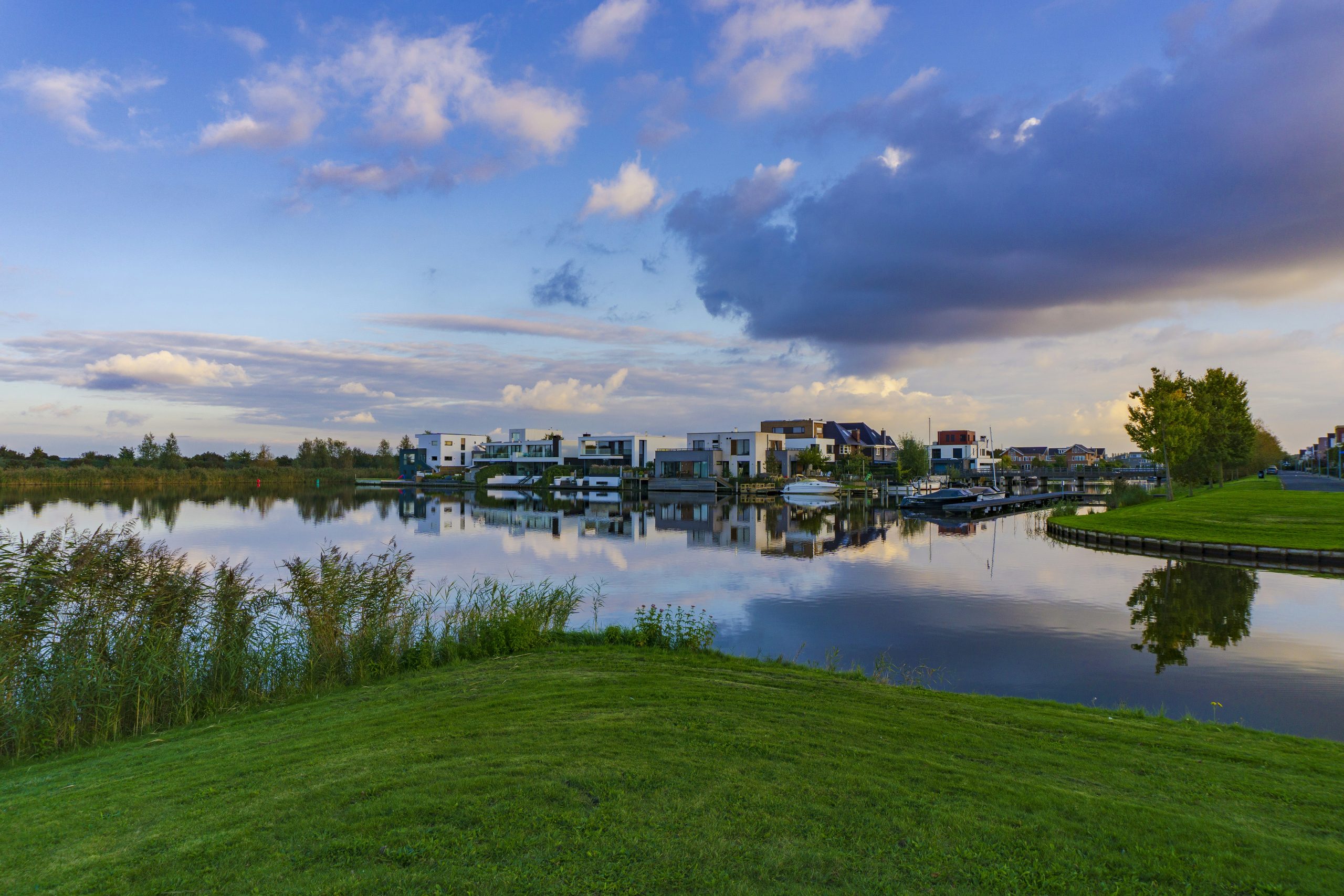 Moderne woningen aan rustig water met duidelijke reflecties, omringd door gras en riet onder een zachte lucht.