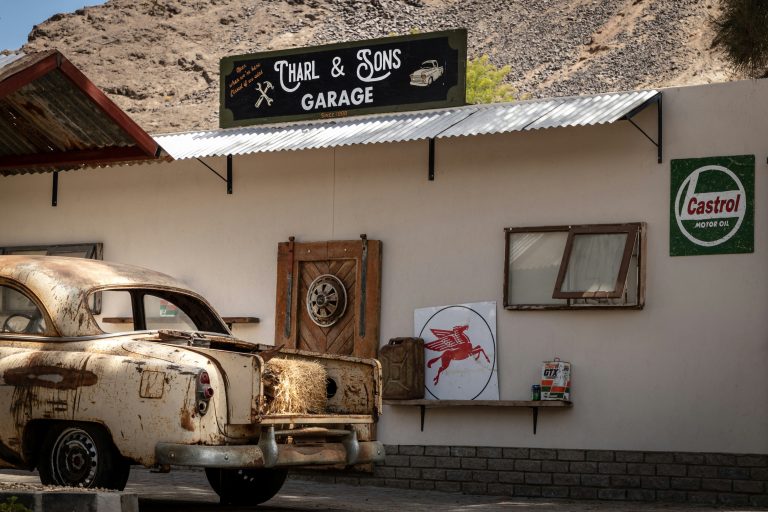 Oude roestige auto geparkeerd naast een kleine garage met vintage borden en gereedschap, in een droog en rotsachtig gebied.