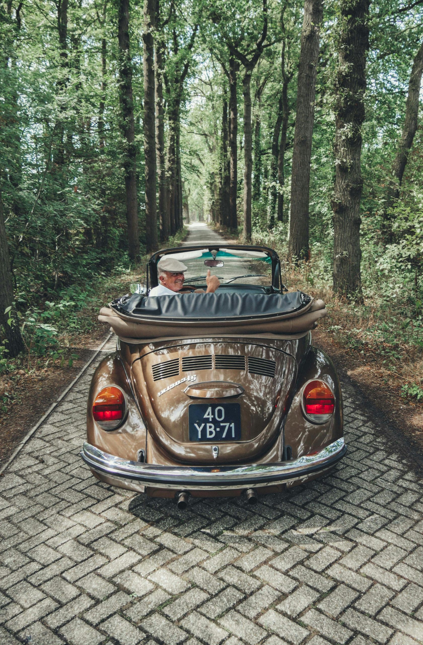 Bruine Volkswagen Kever cabrio rijdend over een smalle bomenlaan, bestuurder met witte pet steekt duim op.